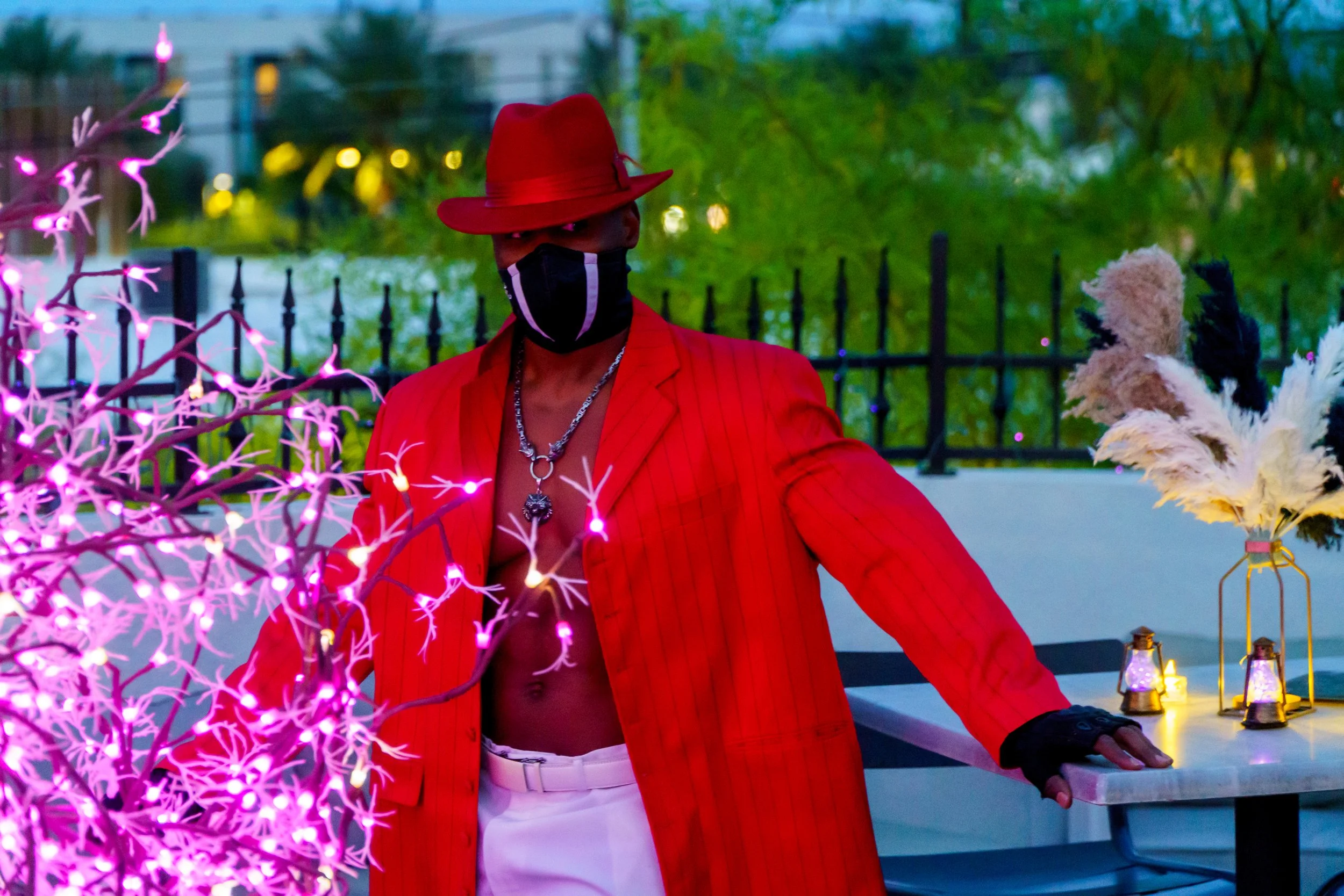 Person wearing a red hat, black face mask, red blazer, and white pants, standing outdoors at dusk near pink illuminated decorative branches and lanterns, with green trees and a black fence in the background.