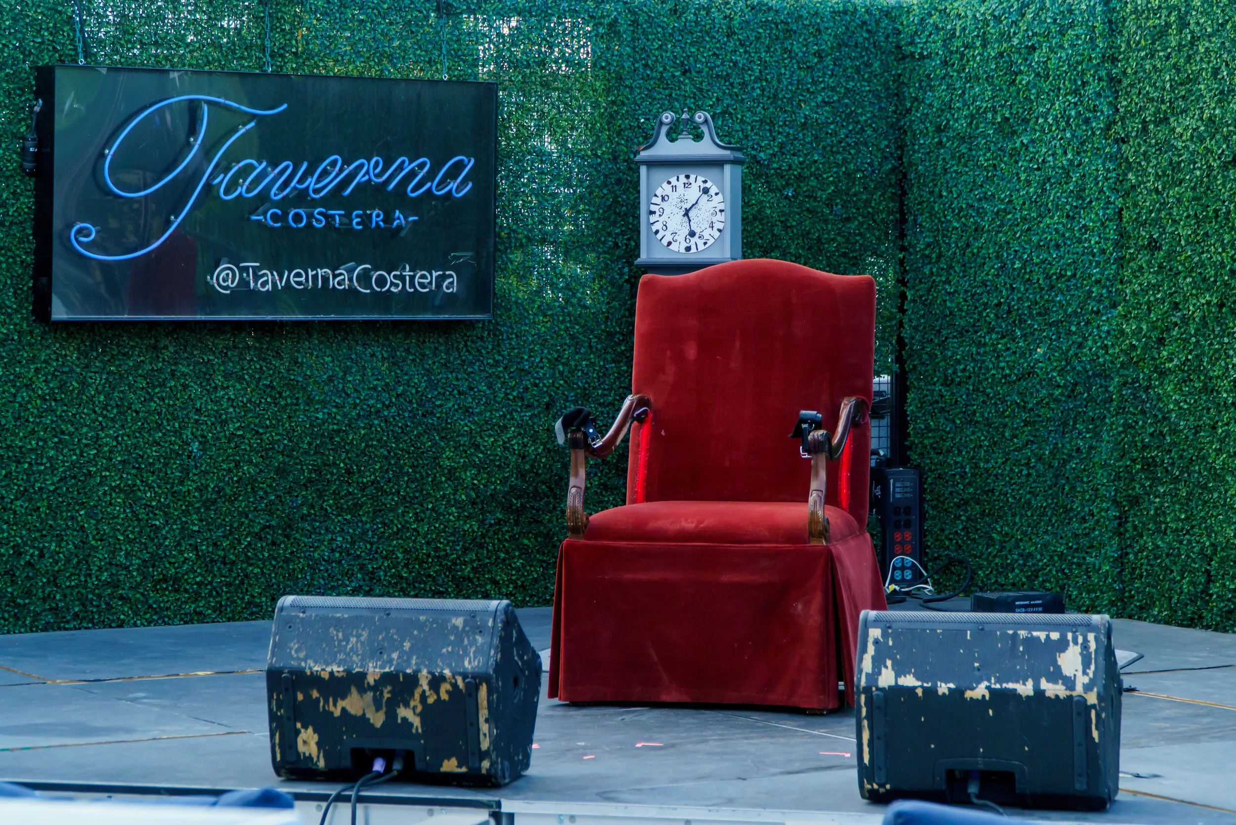 Empty red velvet throne chair on stage with a large clock behind it, set against a backdrop of green foliage, with a neon sign to the left reading 'Taverna Costera' and '@TavernaCostera'.