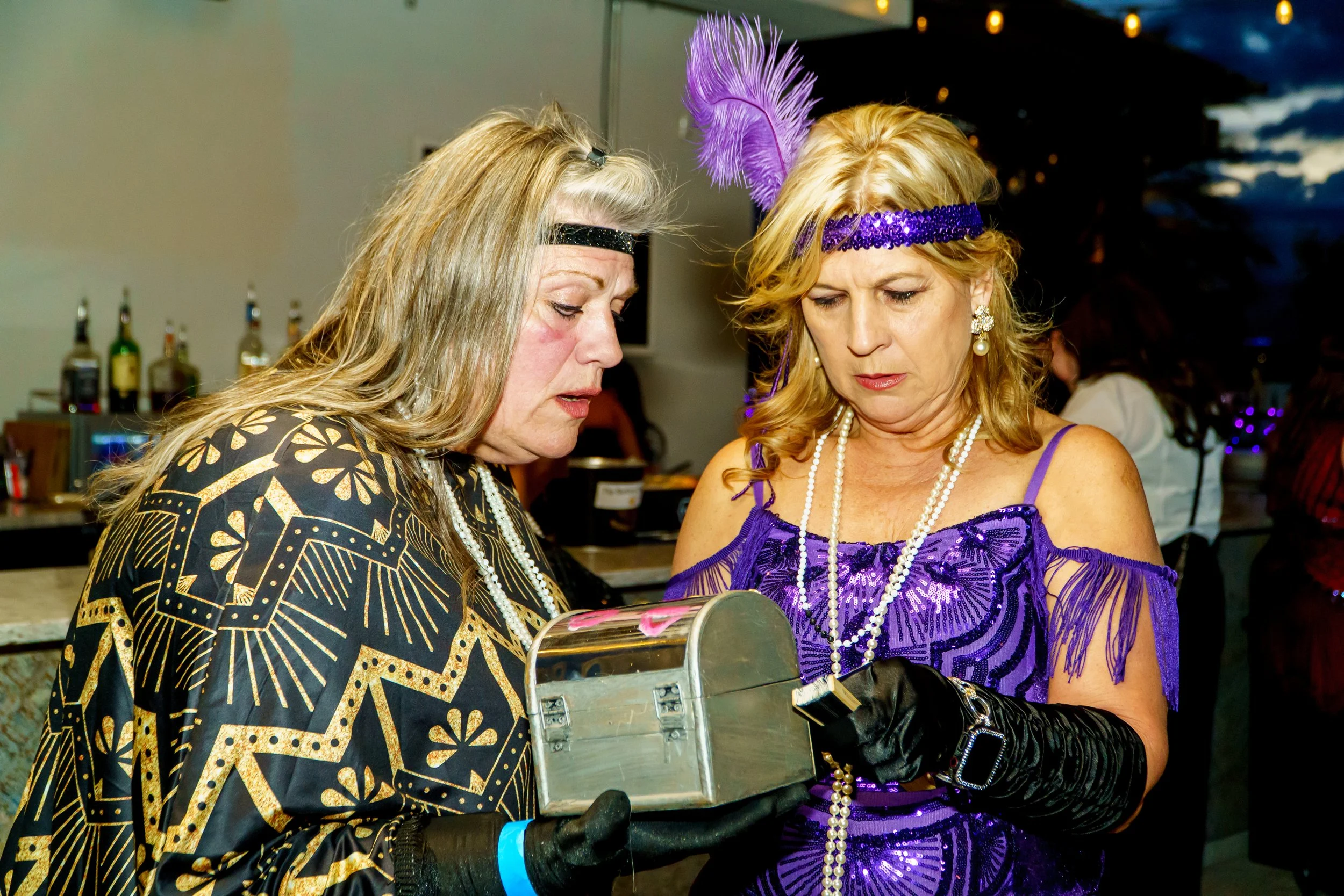 Two women dressed in elaborate costumes with purple feathers, pearls, and accessories look at a cash register at a party or celebration.