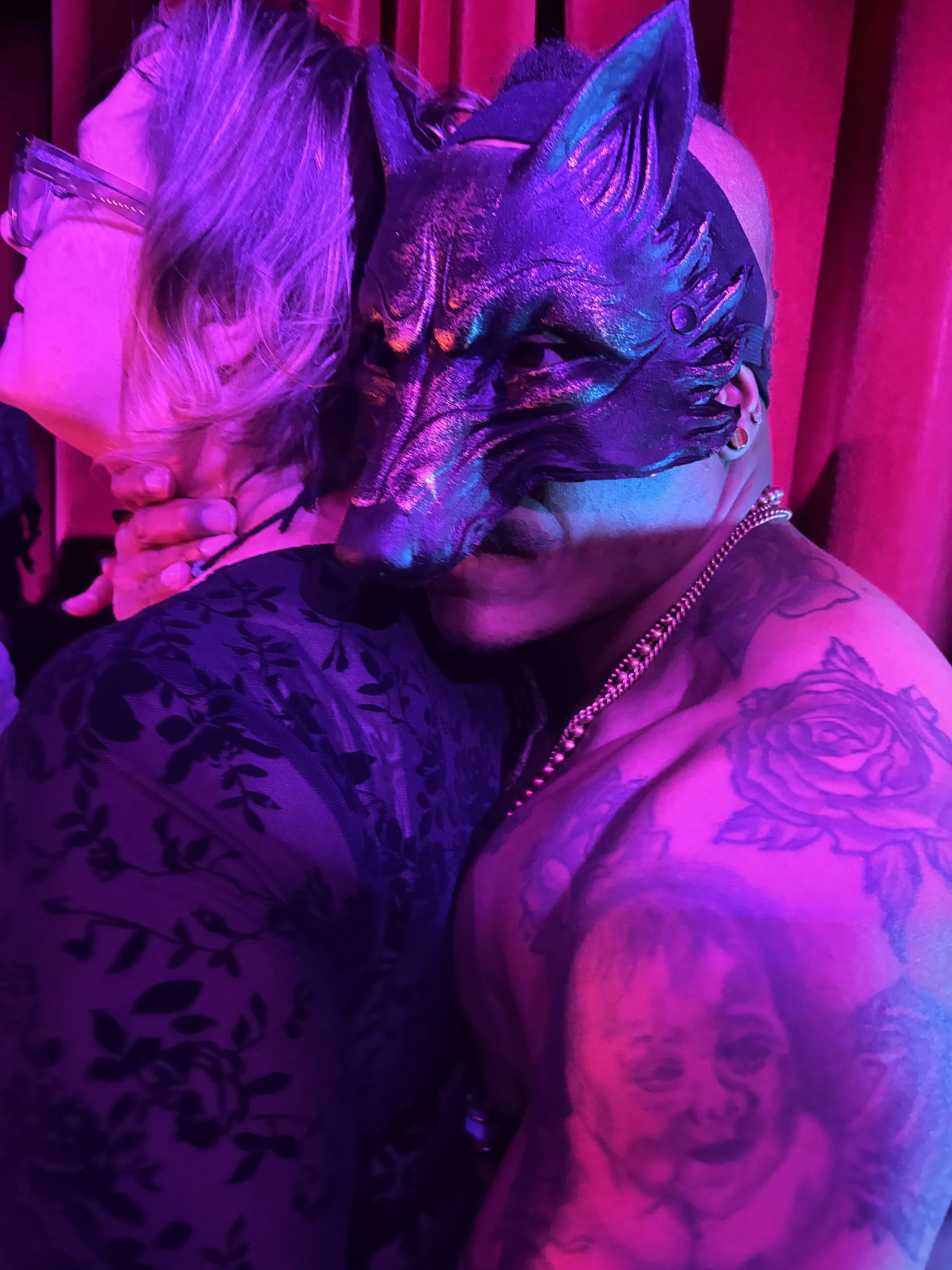 Person wearing a horse mask with a shiny, holographic appearance, illuminated by pink and purple lighting, embracing another person with blonde hair and glasses, in a setting with pink curtains.