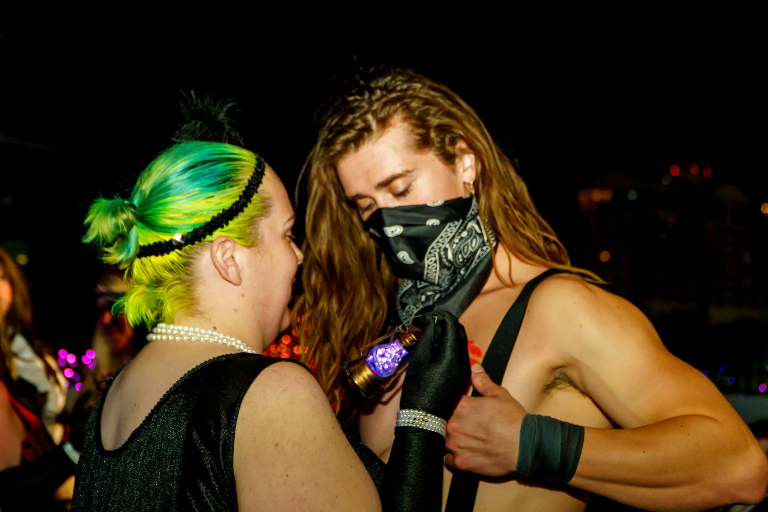 Two women at a nighttime event; one with brightly colored green and yellow hair, pearl necklace, and black top, and the other with curly hair wearing a bandana face mask and black top, embracing and smiling.