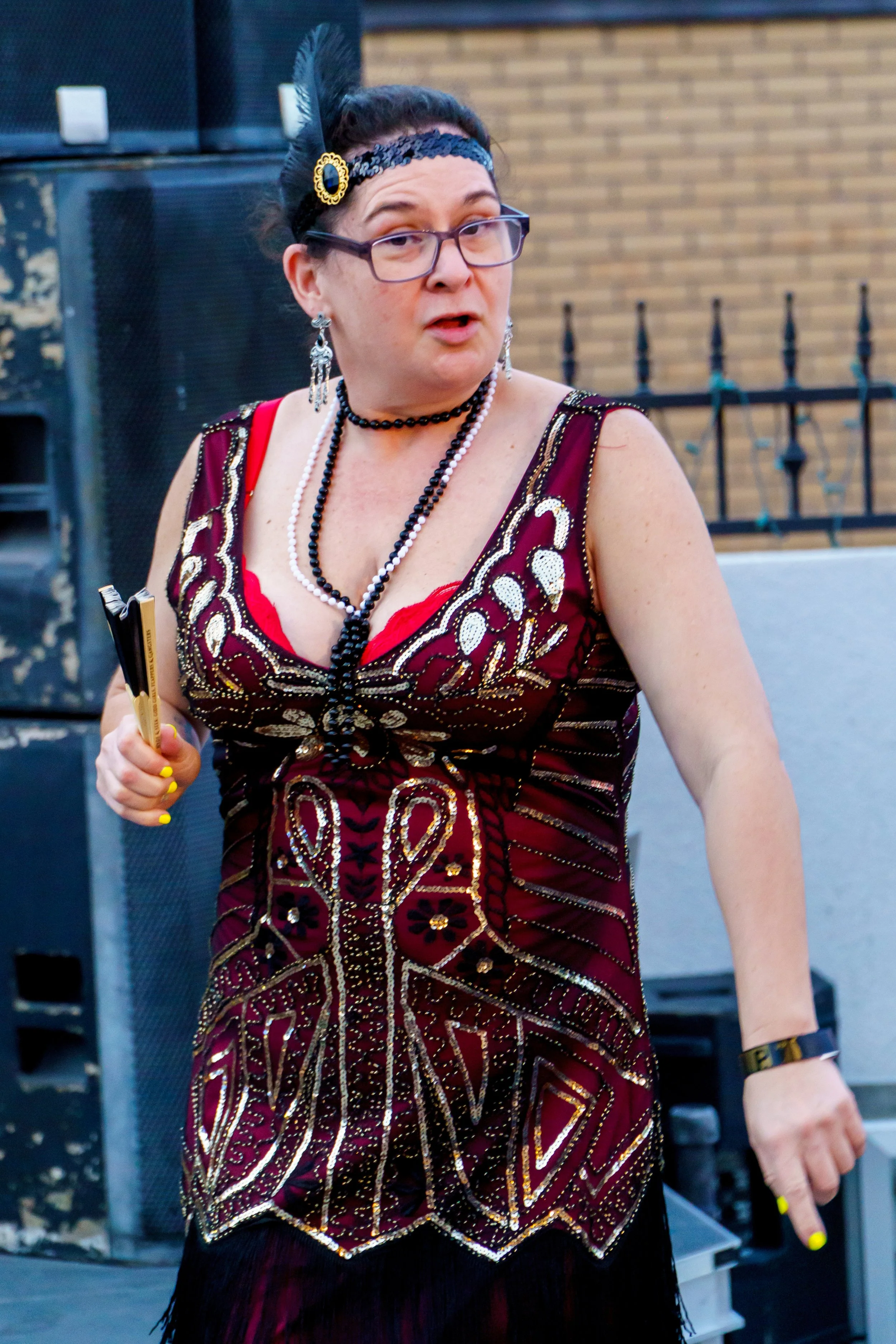 A woman dressed in an elaborate, beaded red and gold dress with matching jewelry, glasses, and a feathered headband, holding a clipboard, standing outdoors with speakers and a brick wall in the background.