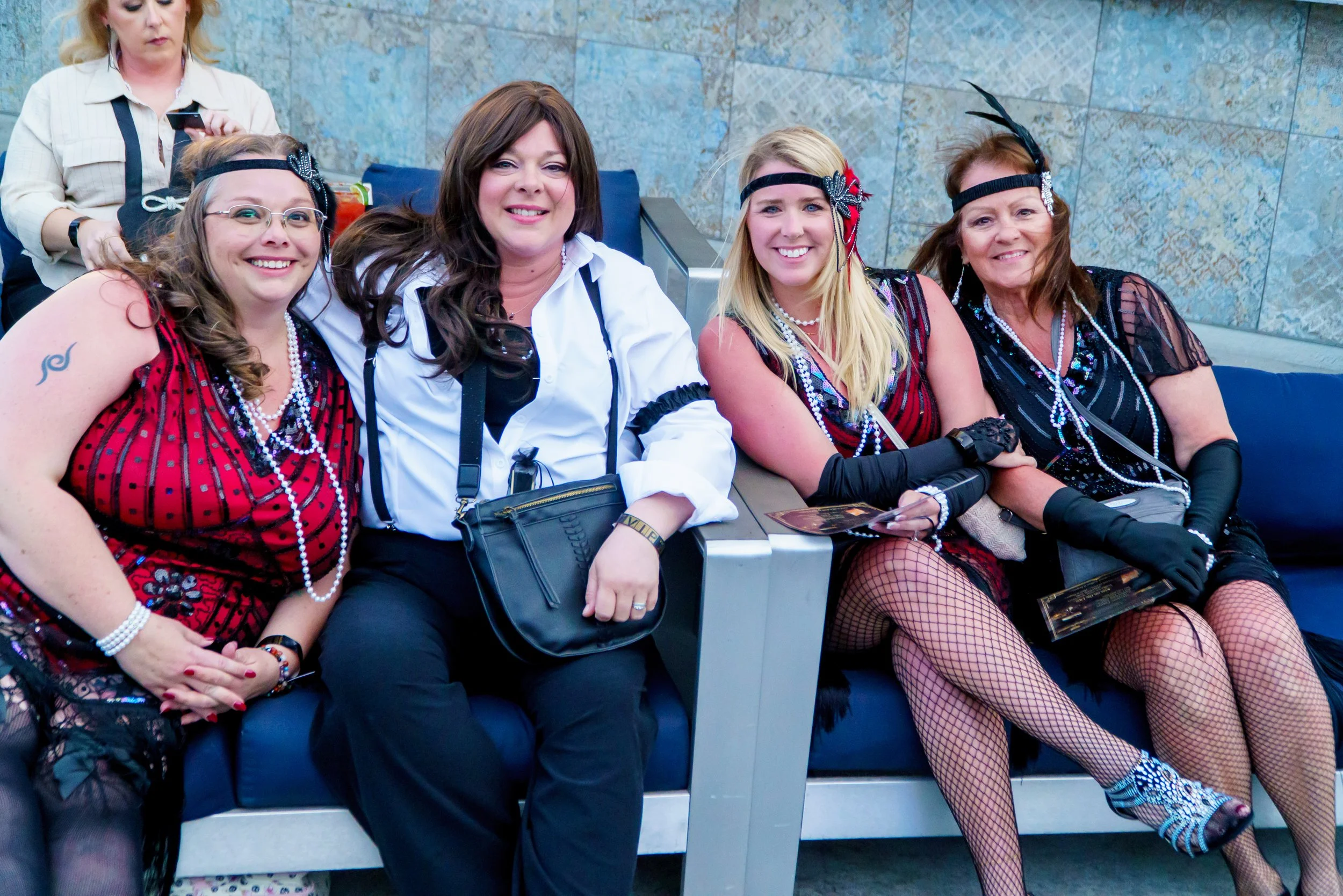 Four women dressed in 1920s flapper costumes sitting together on a bench at an indoor event. They are smiling and wearing accessories such as headbands with feathers, beaded necklaces, and fishnet stockings.