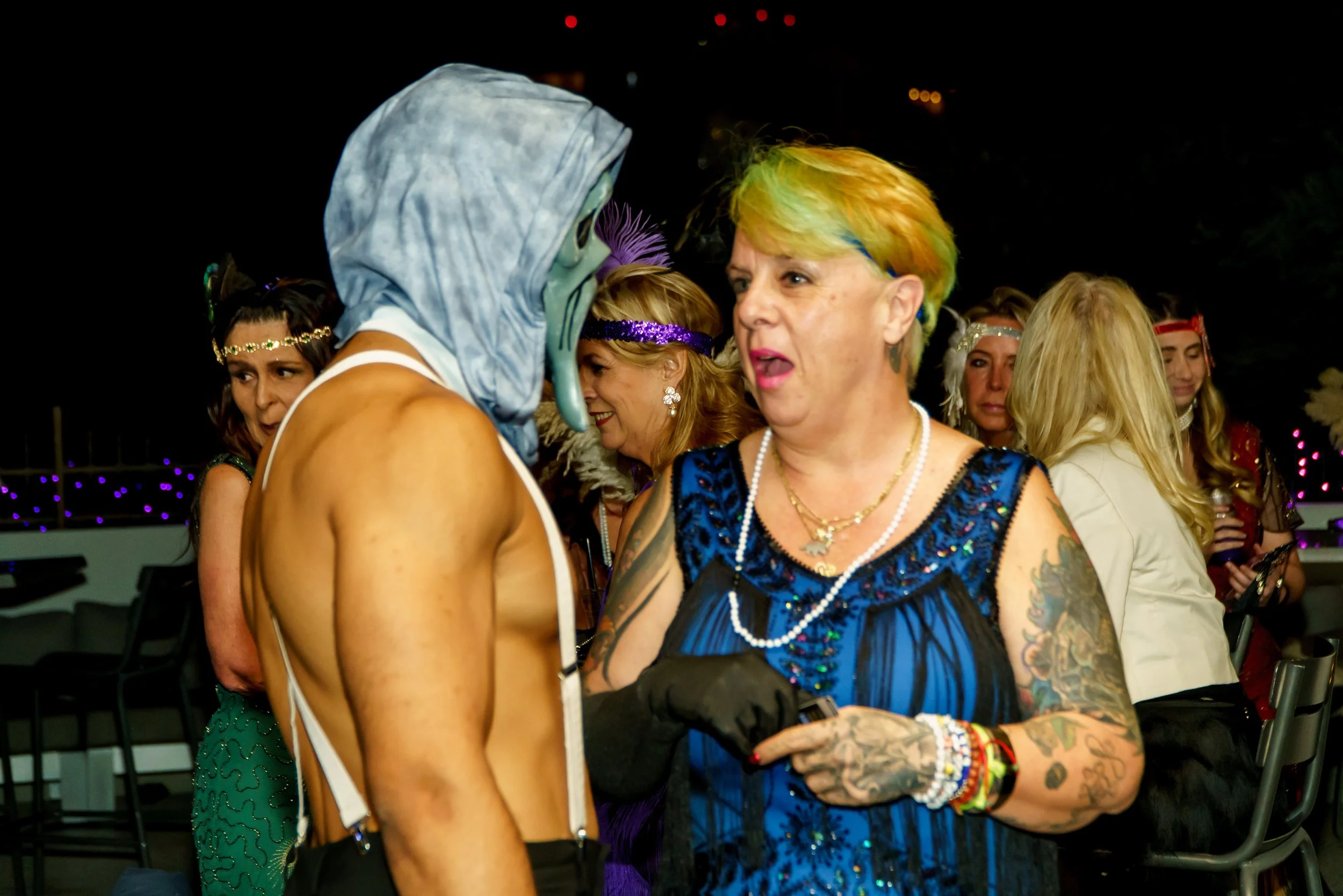 People at a costume party, including a person in a shark costume with a hoodie and mask, and a woman with short rainbow-colored hair, tattoos, and wearing a blue dress, interacting and chatting.