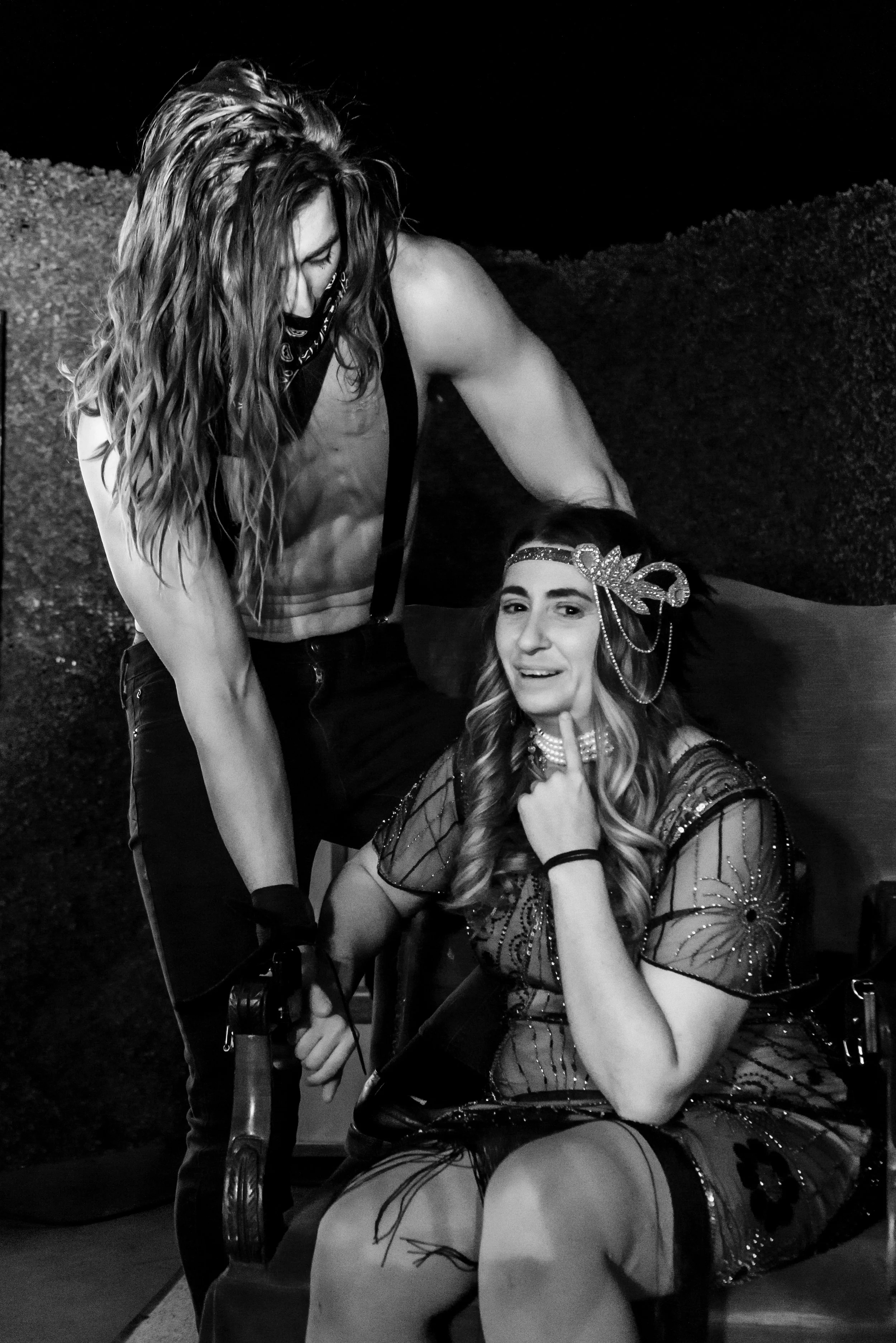 A person with long hair and a bandana riding a bicycle at night, another woman with a decorative headpiece and patterned dress sitting on a chair, smiling and pointing upward.