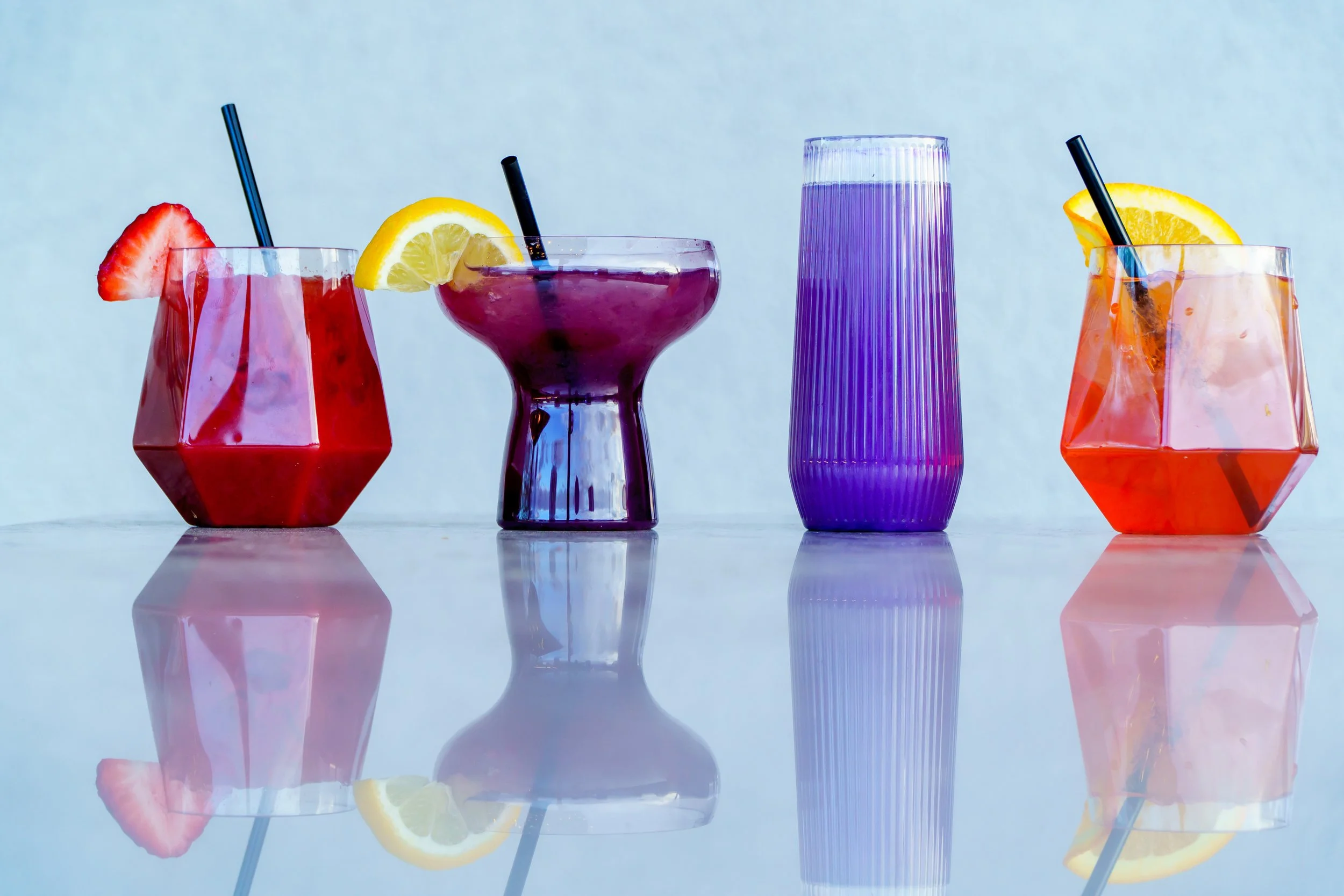 Four colorful glasses of beverages with fruit garnishes, placed on a reflective surface with a light blue background.