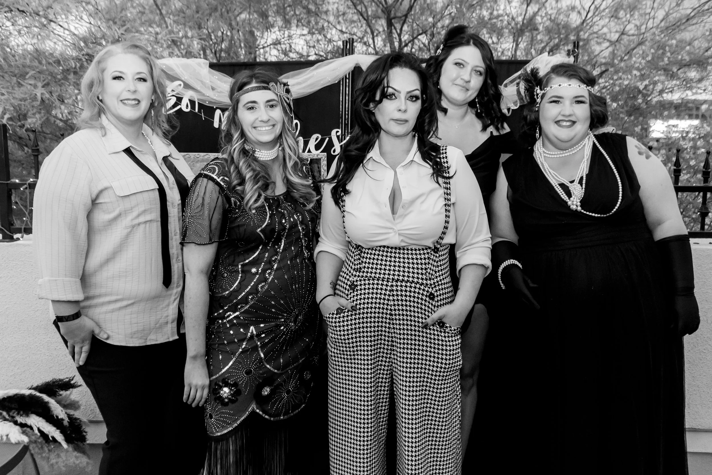 Five women are posing outdoors for a group photo. They are dressed in vintage or retro style clothing and accessories, with some wearing pearl necklaces and headbands. The background shows leafless trees and a dark fence with a decorative sign.