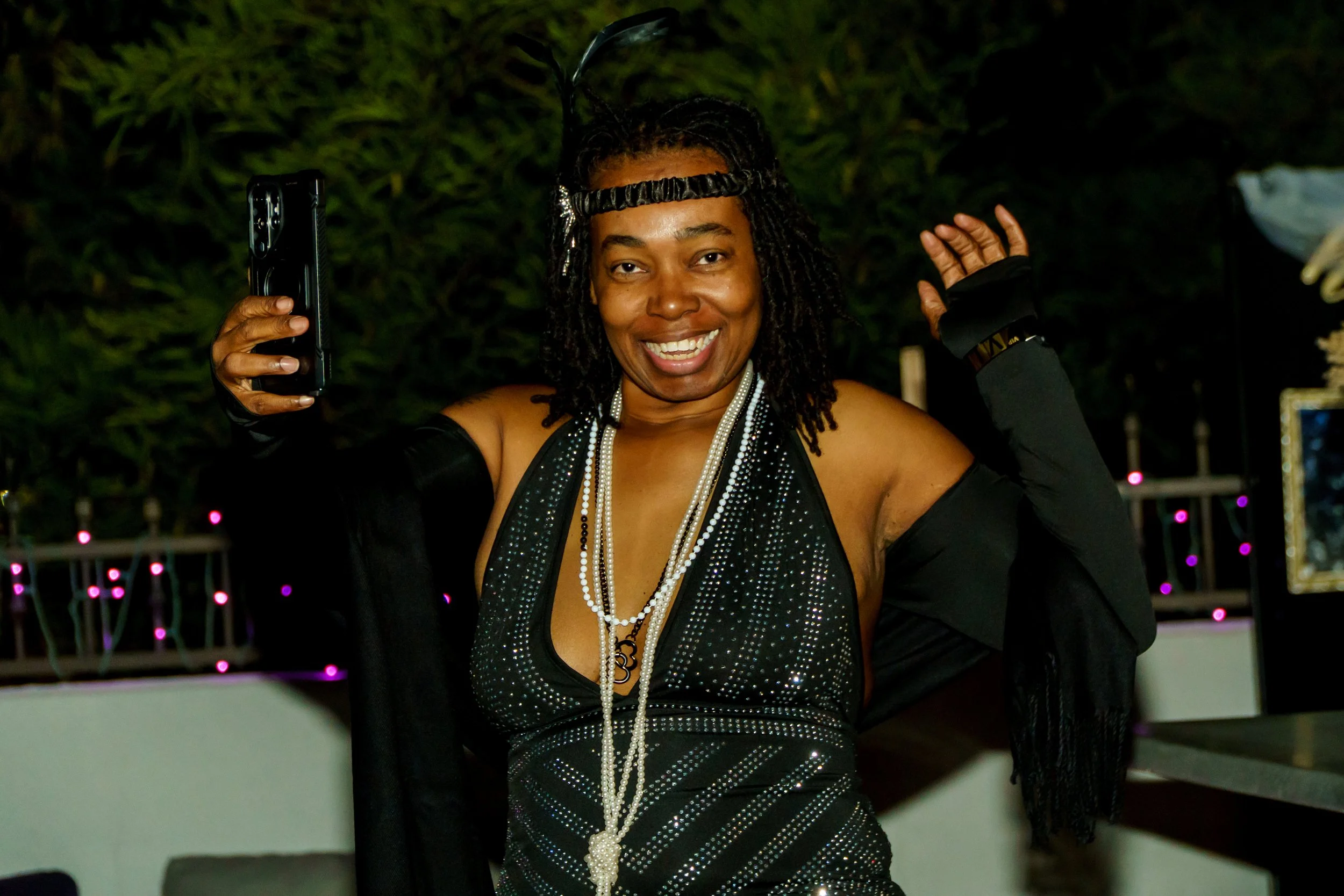 A woman in a black sequined dress smiling, wearing pearl necklaces and a headband with a feather, at night with decorative lights in the background.