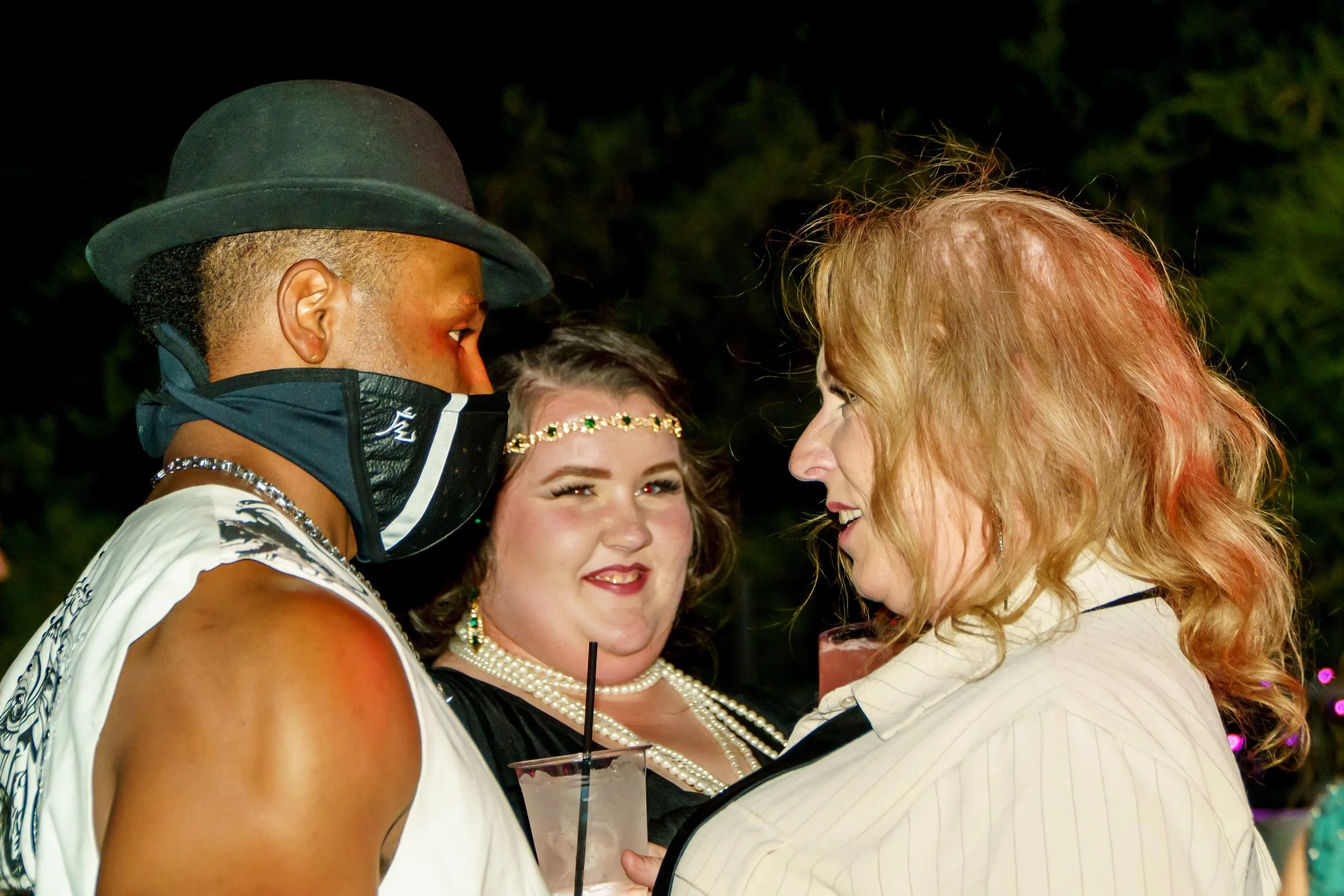 Four women are standing close together, smiling and engaging in conversation, at night with blurred lights in the background.