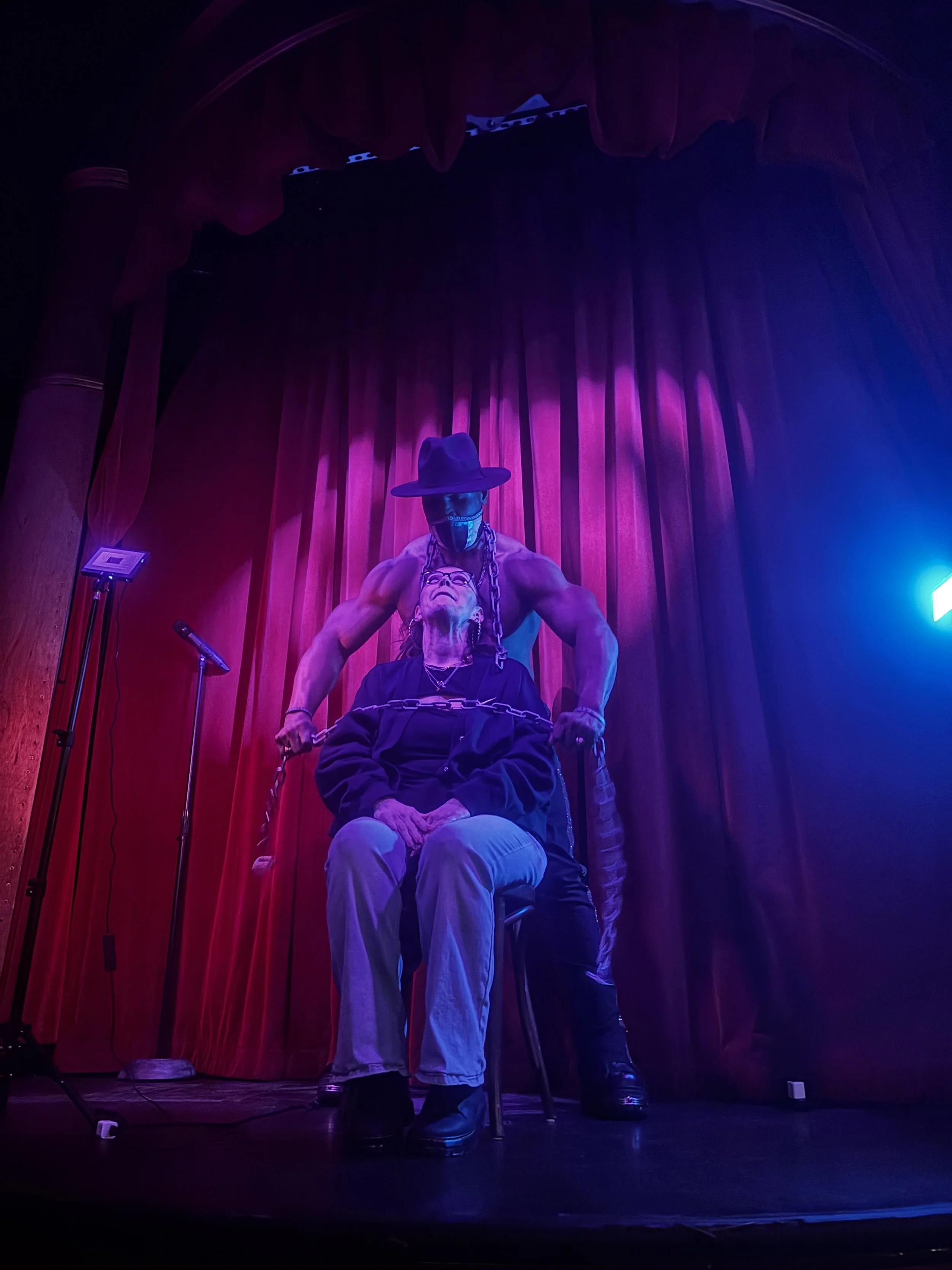 A stage with red curtains and purple lighting featuring a muscular man in a hat and chain, holding an unconscious or restrained woman sitting in a chair.