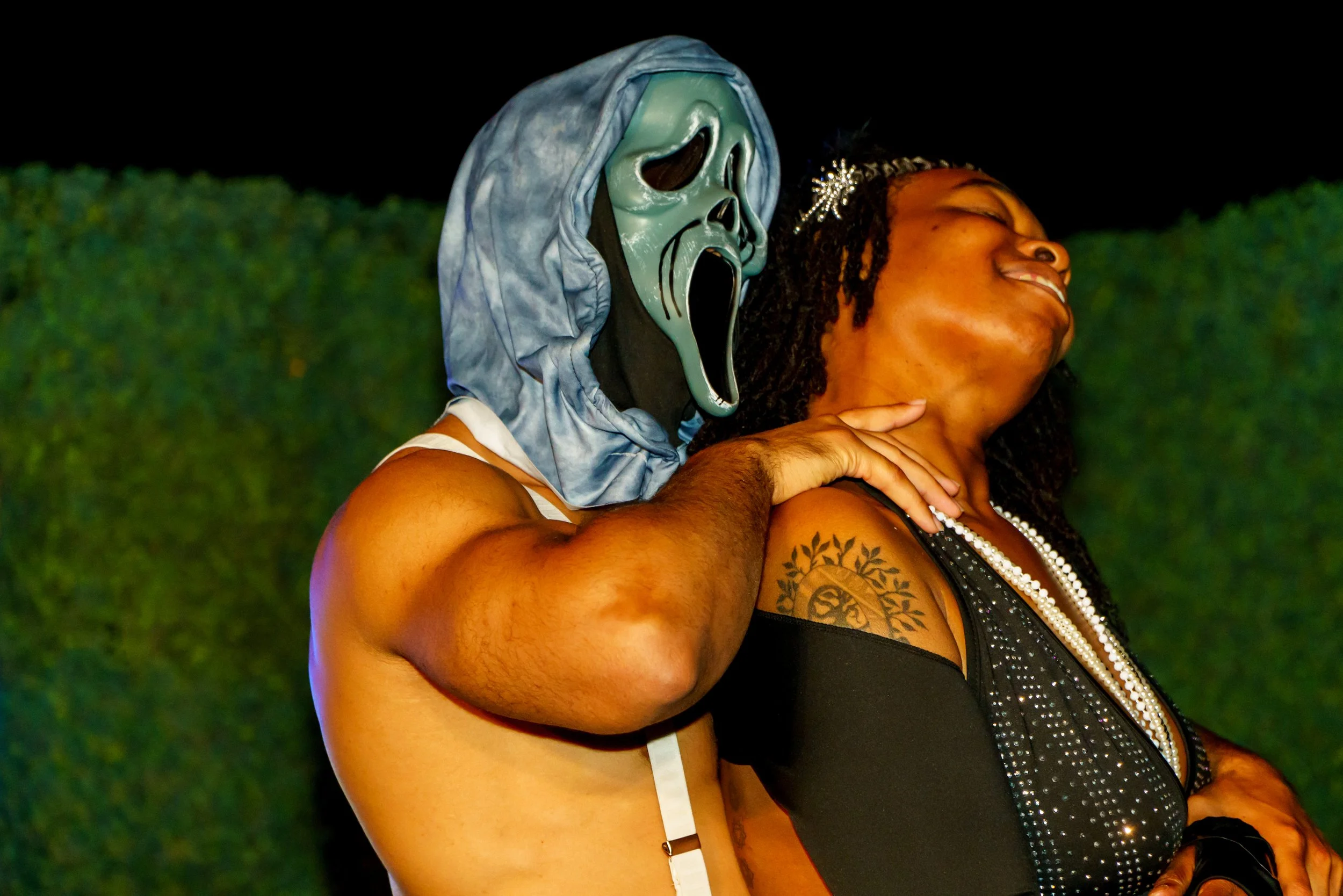 A woman with tattoos on her arm and dressed in black with a pearl necklace is smiling with her eyes closed while leaning her head back. Another person, wearing a mask with a skeleton face and a hood, is behind her and appears to be holding her around