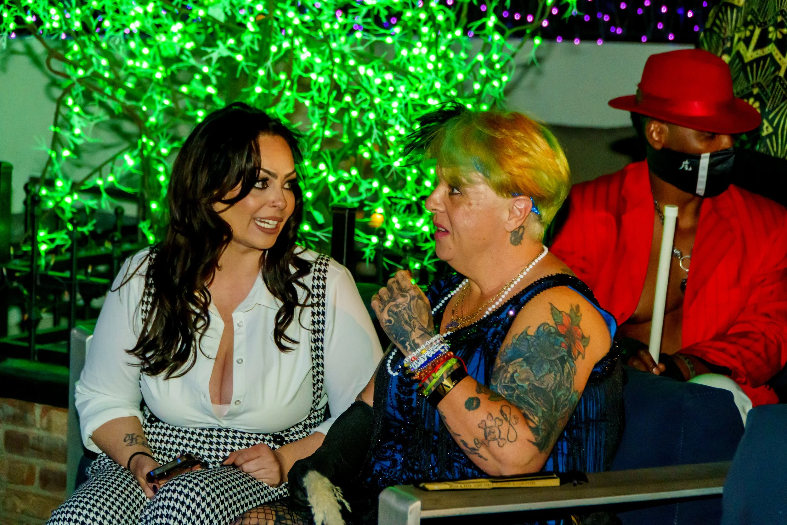 Two women are sitting close together, engaged in conversation, with a person wearing a red hat and mask sitting nearby. The background features vibrant green and purple string lights.