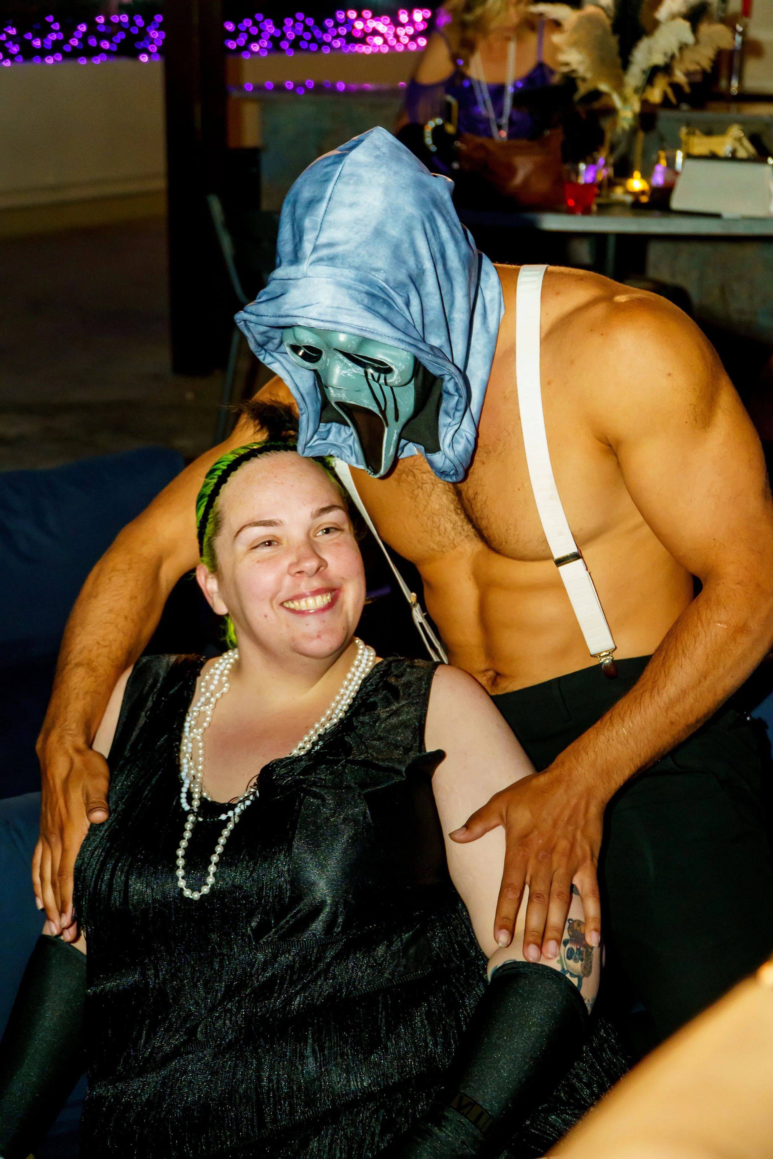 A woman with a pearl necklace and black dress smiling, while a shirtless man wearing a blue mask and hoodie bends over her at a party.