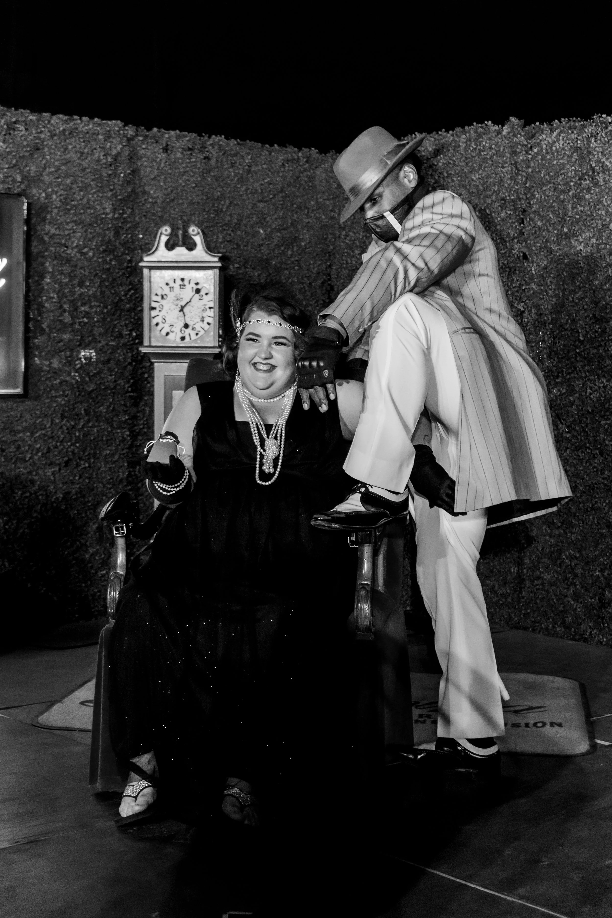 A woman in a wheelchair wearing a black dress and pearl jewelry is smiling and holding a mobile phone. A man in a striped suit, wide-brimmed hat, mask, and gloves is standing next to her, wearing a pinstripe suit, and leaning towards her in a pose. T