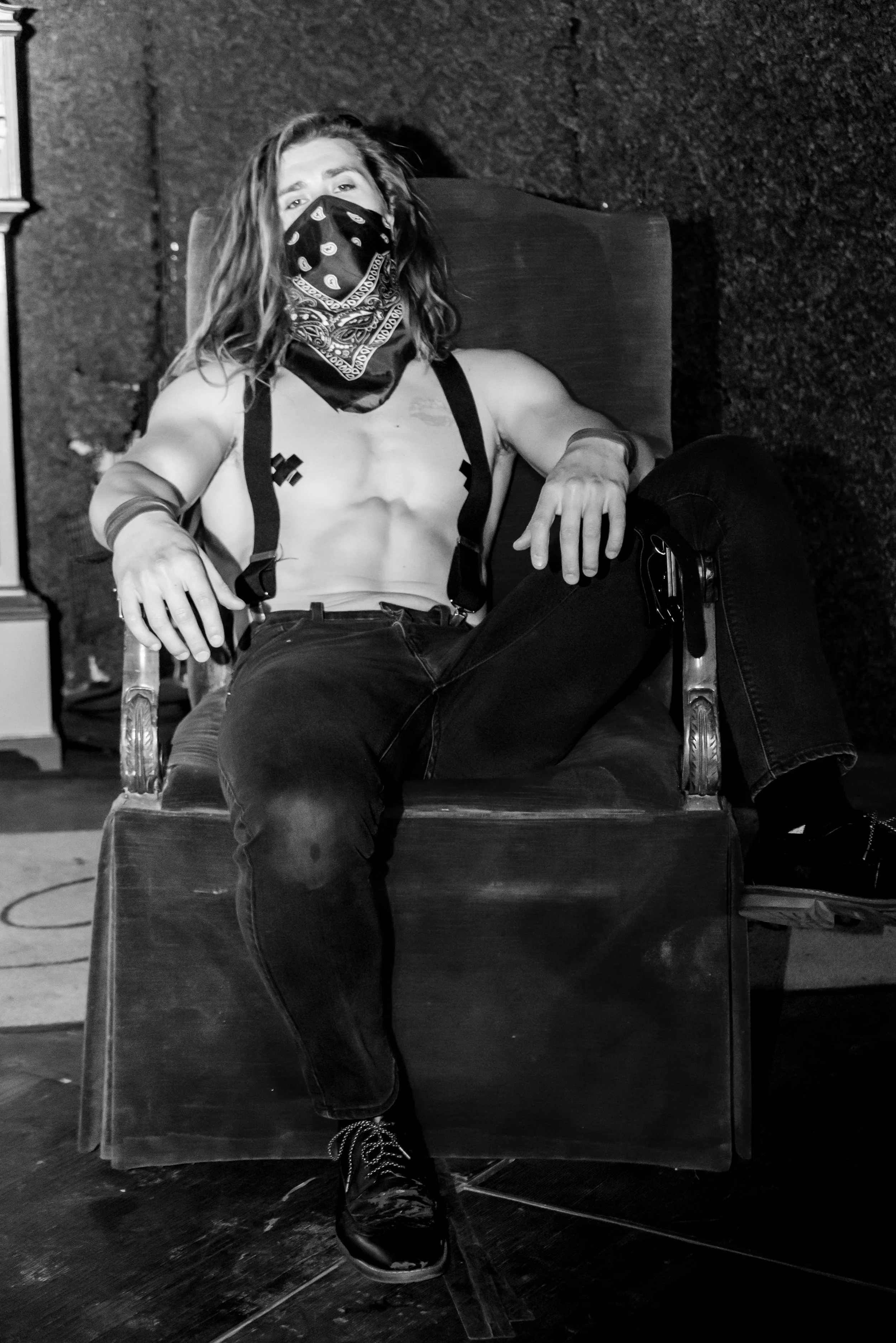 A person with long hair, wearing a bandana over their face, sitting in a chair in a dimly lit room with textured walls, dressed in dark clothes, with their shirt partly open showing their chest and tattoos. They are looking at the camera, with one ar