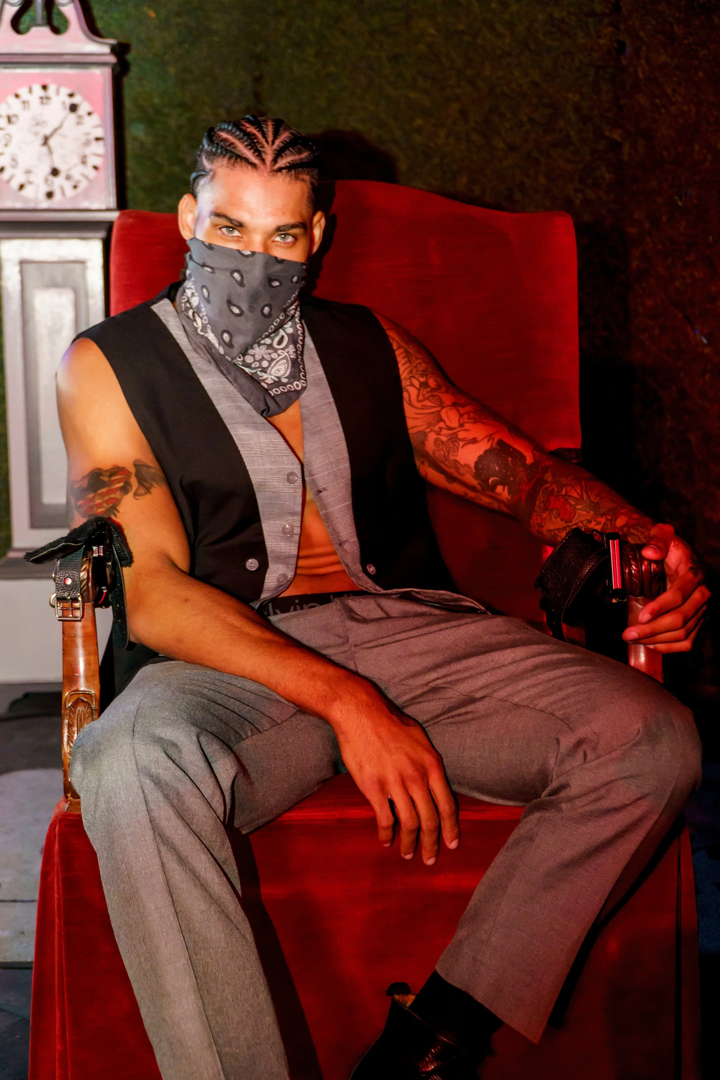 Young man with braided hair and tattoos sitting on a red velvet chair wearing a bandana mask, gray suit pants, sleeveless vest, and black shoes, holding a water bottle, with a clock in the background.