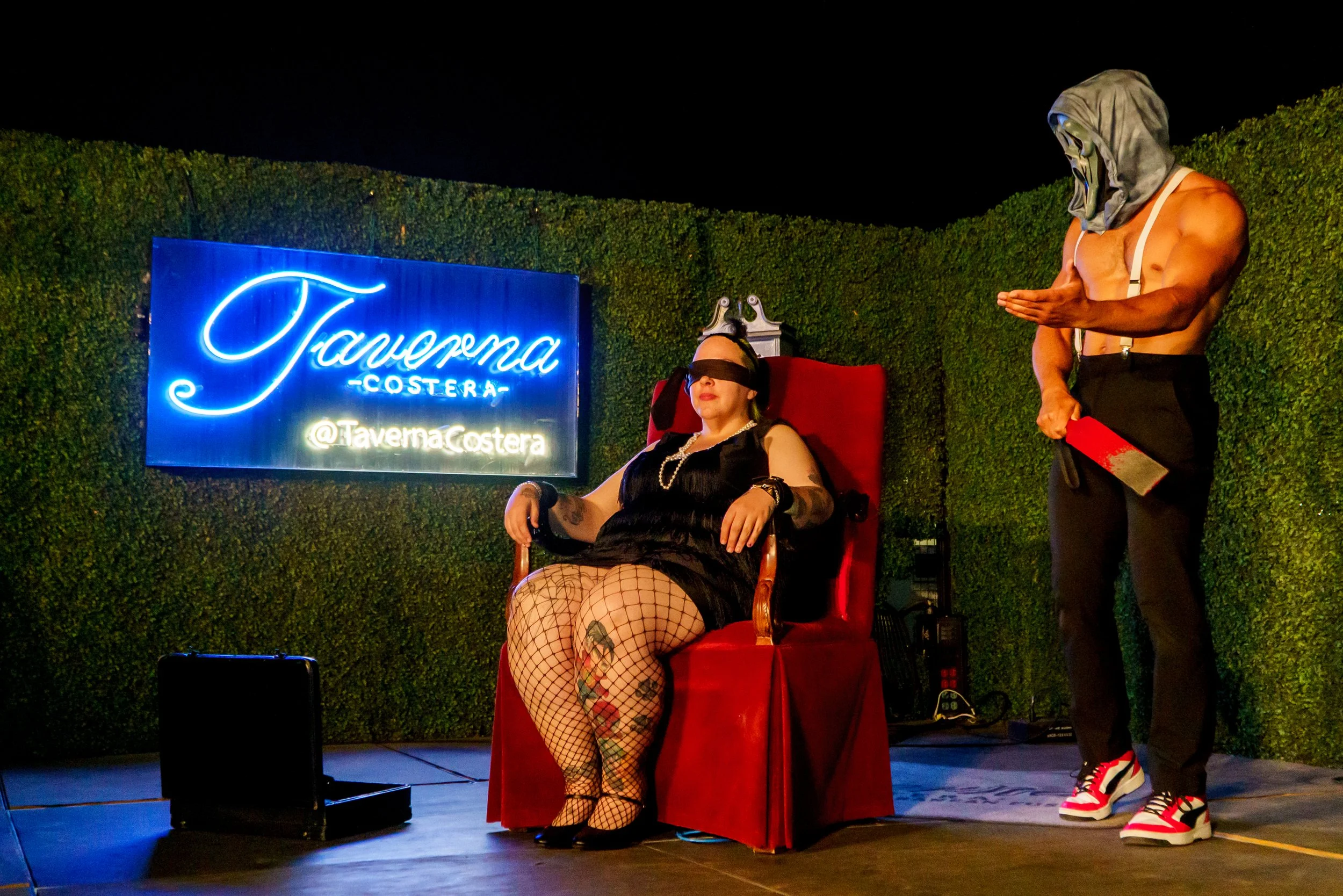 Woman with tattoos and fishnet stockings sitting in a red chair, wearing a blindfold and black dress, next to a man with a hood and mask holding a cleaver. They are outside at night with a hedge and a neon sign that says 'Taverna Costera'