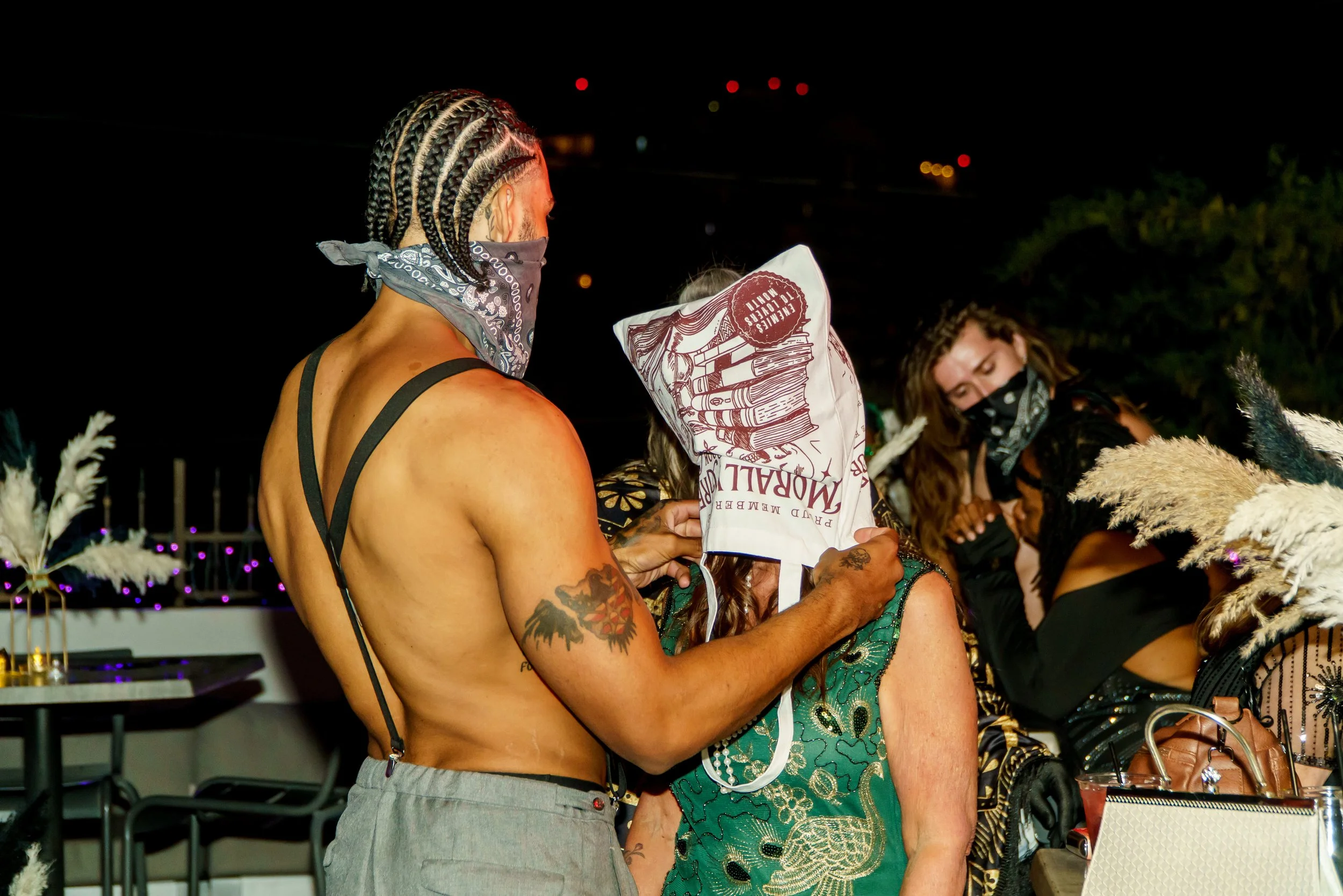 A shirtless man with braided hair and tattoos on his arm holding a newspaper while interacting with an older woman, with other people in the background at night.