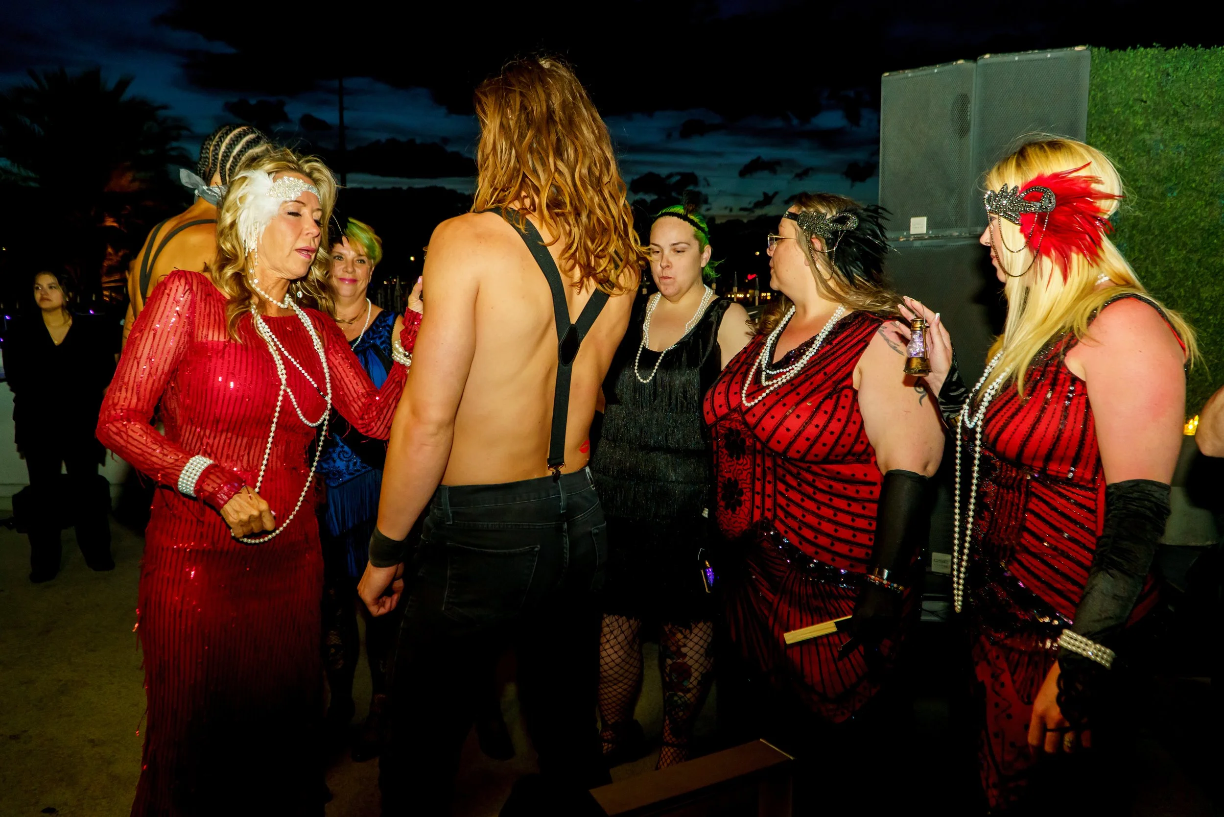 Group of women dressed in cabaret or burlesque costumes, some wearing feathered headdresses with sequins, gathered around a shirtless man with long red hair during an outdoor night event.