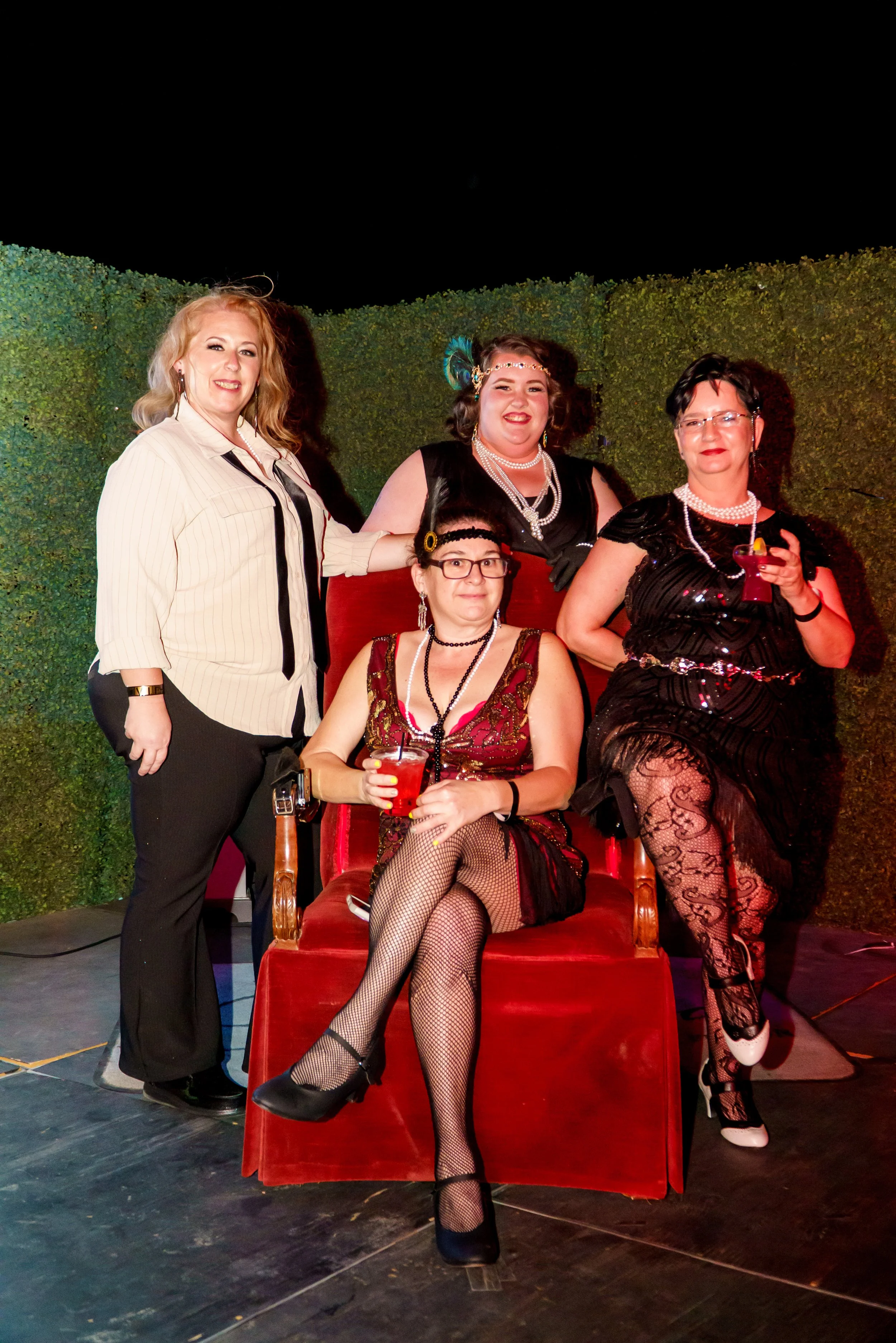 Four women dressed in vintage or retro clothing, with the one seated in front holding a drink,most likely at a themed party or event, with a green hedge wall behind them.