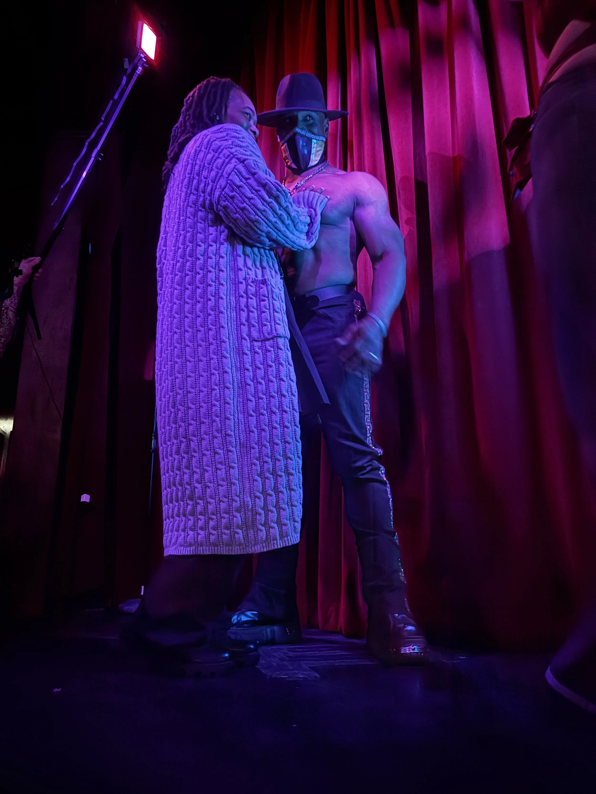 Two people on a stage in front of red curtains, one shirtless with a hat and mask, the other wearing a long knitted coat, under purple lighting.