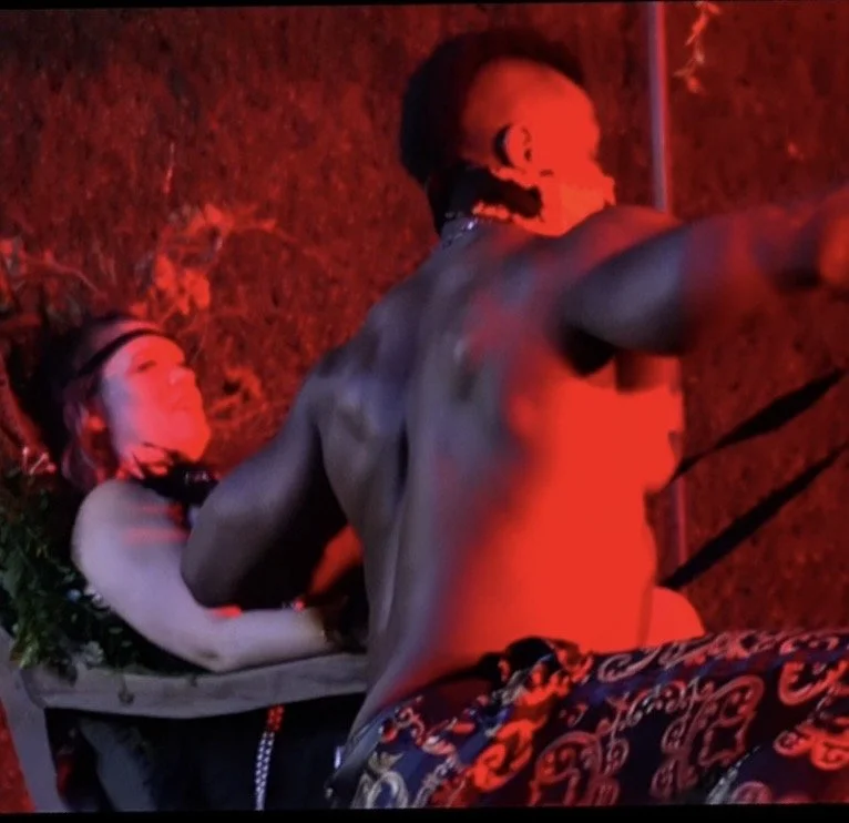Two individuals, one shirtless male with tattoos and necklaces, and one female with jewelry and a blouse, are in a dimly lit, red-colored setting, possibly a nightclub or party, engaged in a close interaction.