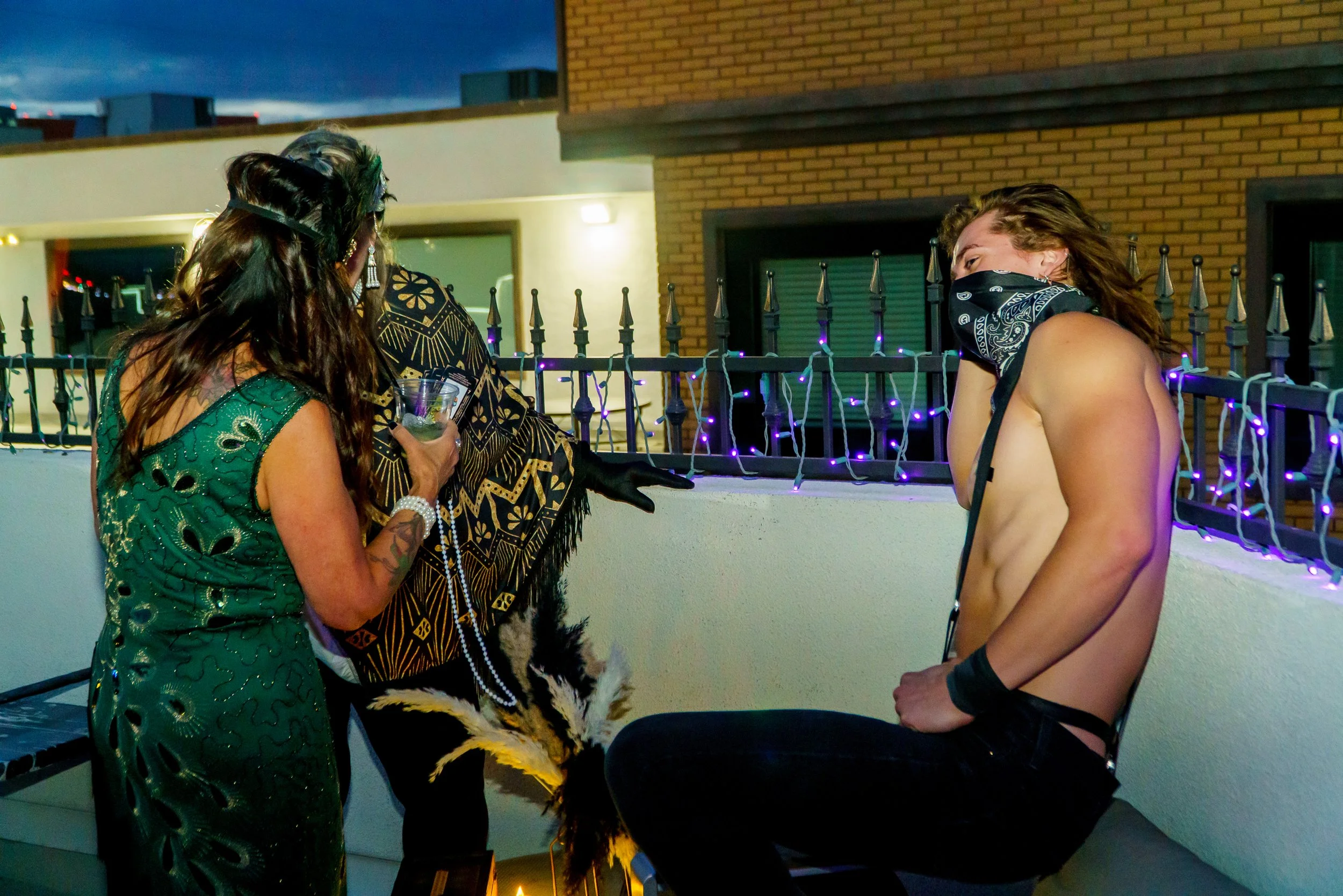 Three people standing outdoors on a balcony at dusk, one woman with long dark hair wearing a green dress, a person in tribal attire with a feathered mask, and a shirtless man with long curly hair and a bandana covering part of his face, engaging in c