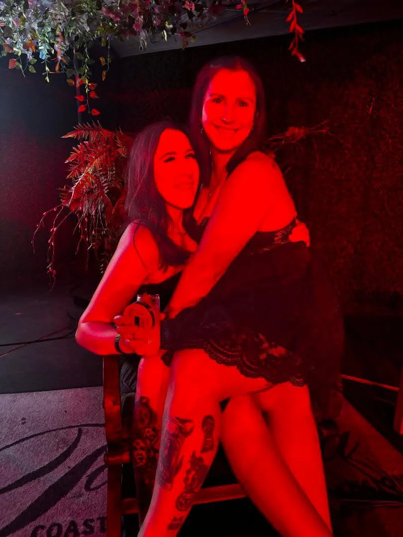 Two women dancing and smiling at a party, illuminated by red lighting, with floral decorations in the background.