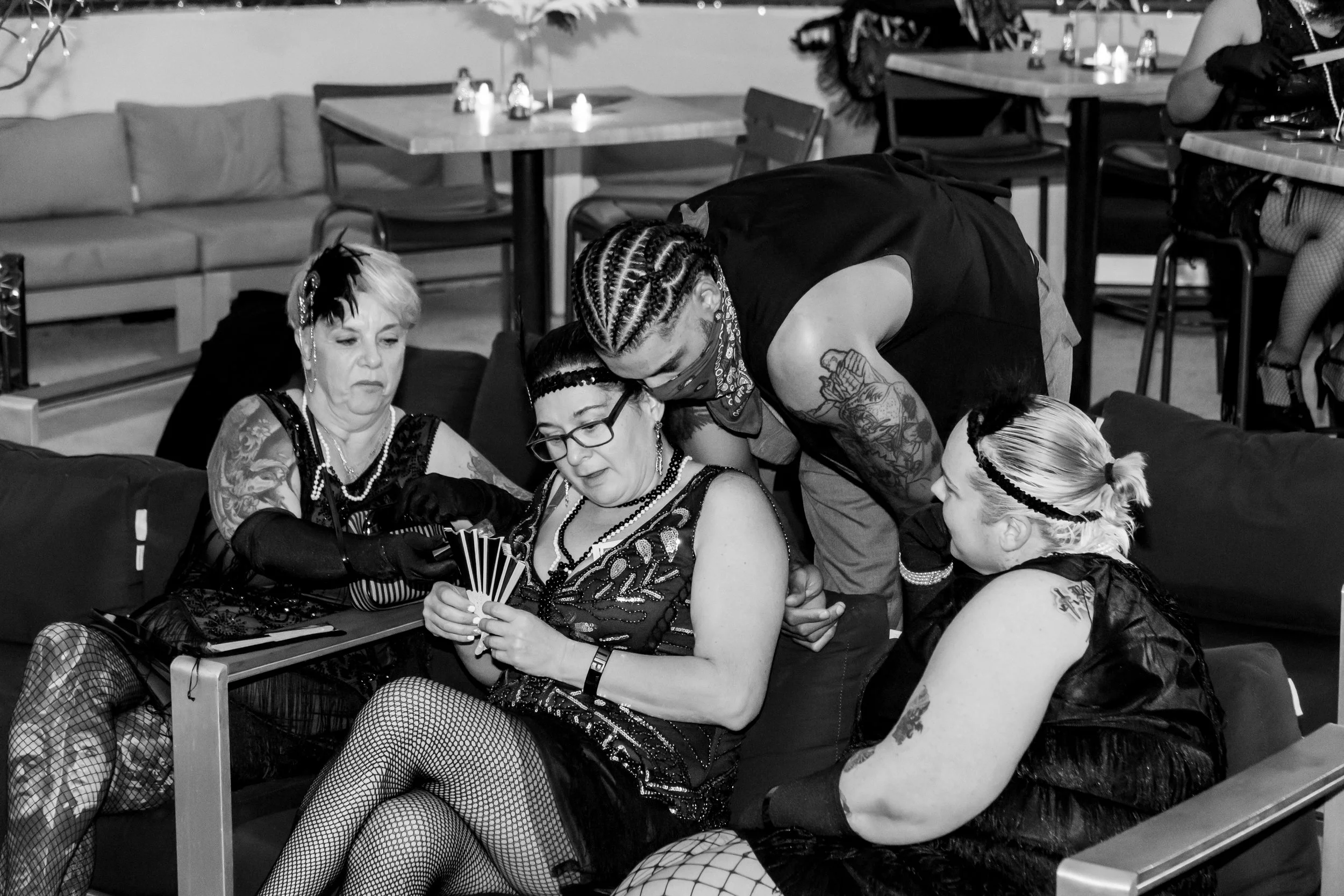 A group of tattooed women dressed in black clothing and fishnet stockings, gathered around a woman holding a fan, in a room with tables and chairs, some lit candles, and decorative plants.