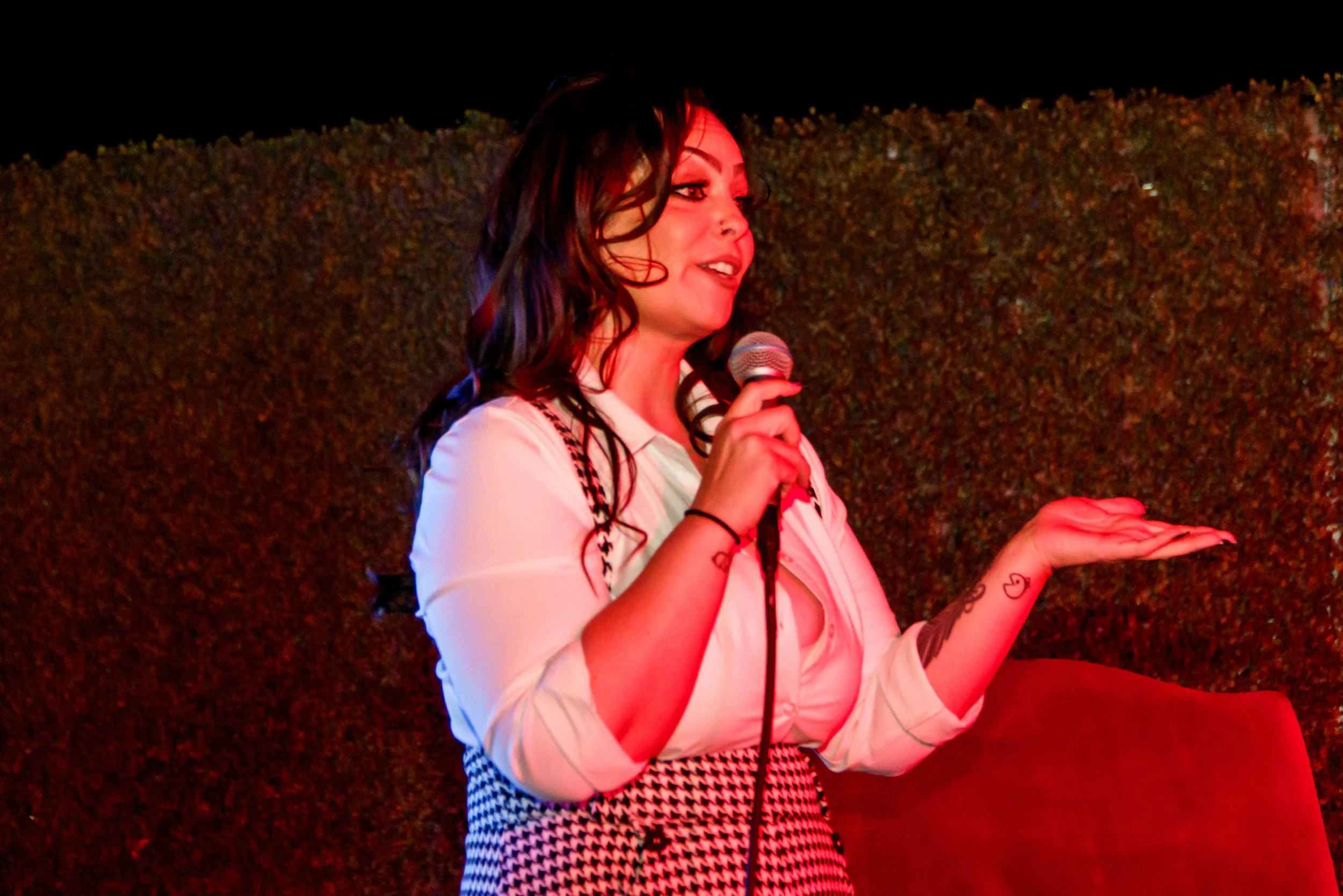 A woman with dark hair, tattoos, and makeup holding a microphone, speaking or performing in front of a textured background.