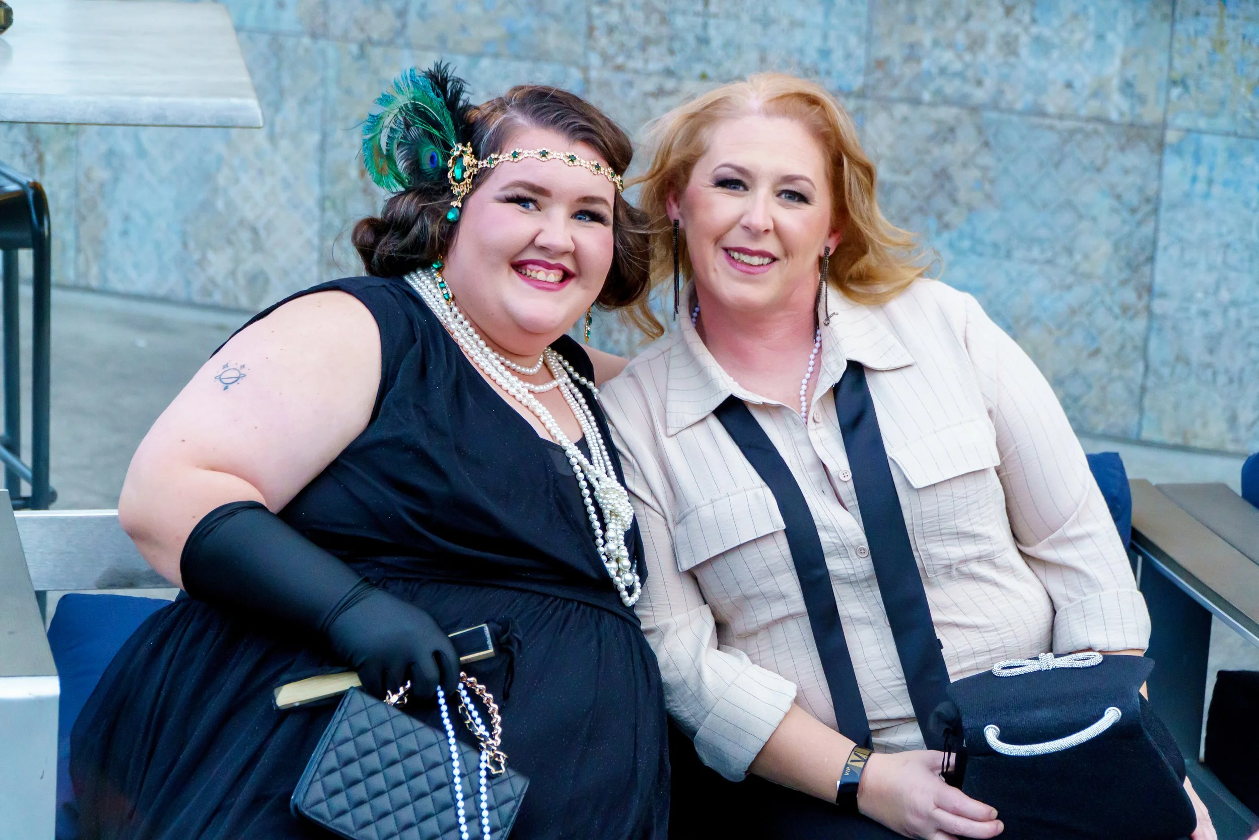 Two women sit side by side on a bench, smiling at the camera. The woman on the left has dark hair, is wearing a peacock feather headband, pearl necklaces, and a black dress. The woman on the right has red hair, is wearing a light-colored striped shir