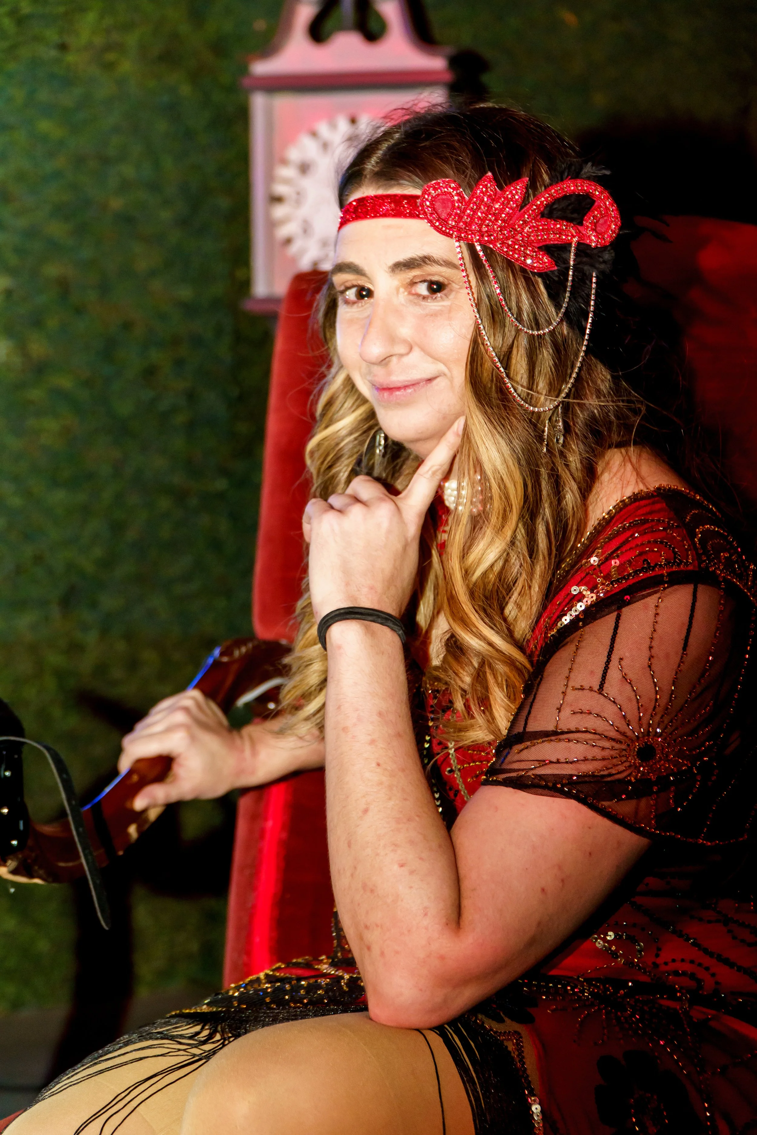 Woman with a red glittery headband, wearing a black and red dress with sequins and mesh, sitting on a red velvet chair, with a green background and a decorative clock in the background.