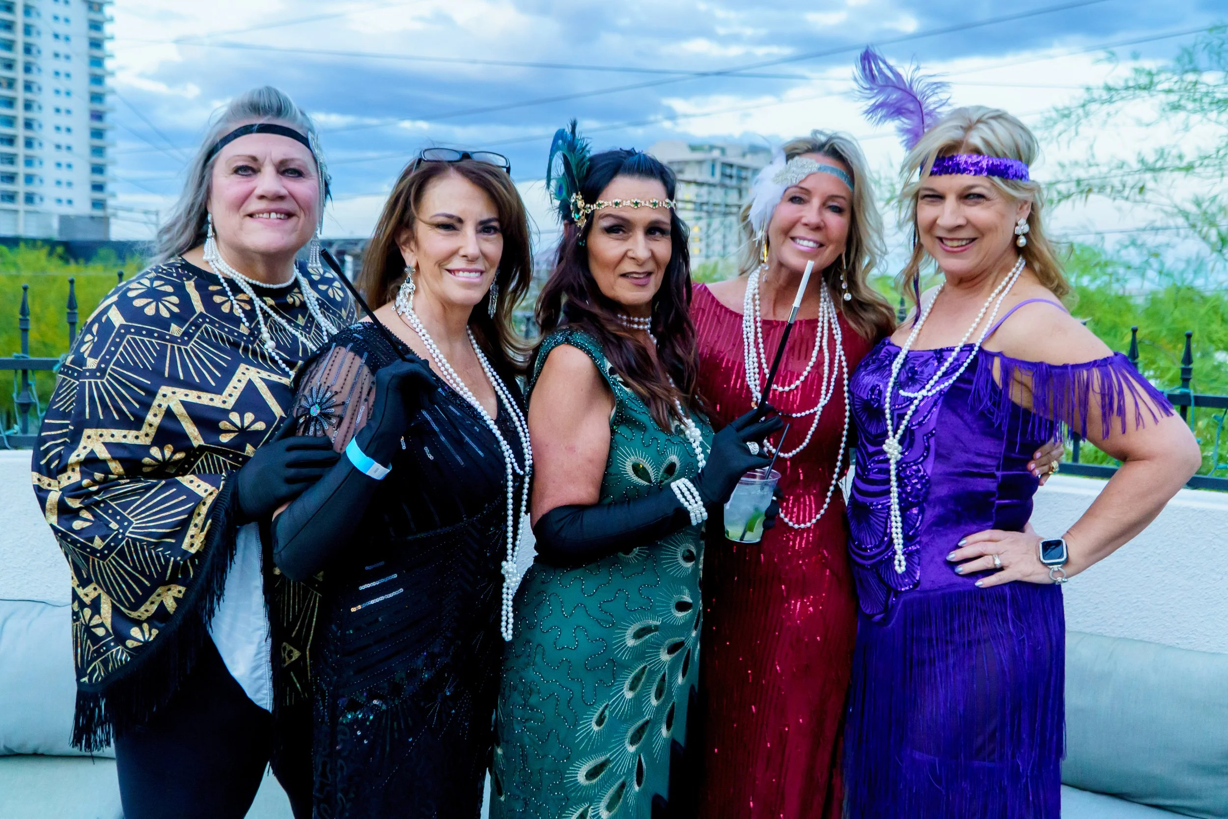 Six women dressed in colorful, vintage 1920s flapper costumes with feathered headbands and pearl necklaces, standing outdoors on a city street with buildings and trees in the background.