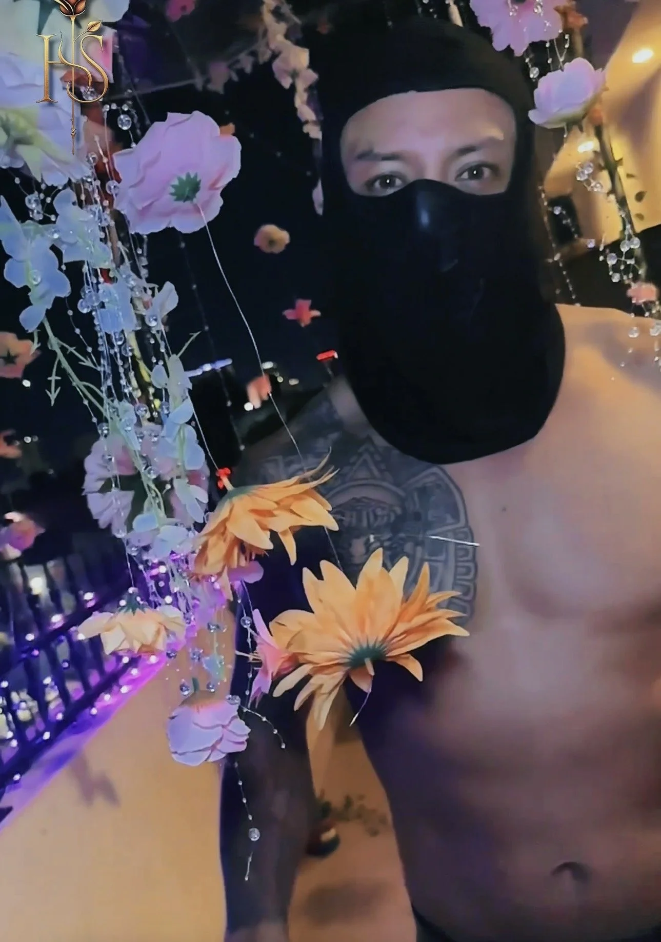 A person wearing a black mask and black clothing, surrounded by colorful artificial flowers and jewelry, with a decorative background filled with purple and yellow lighting.