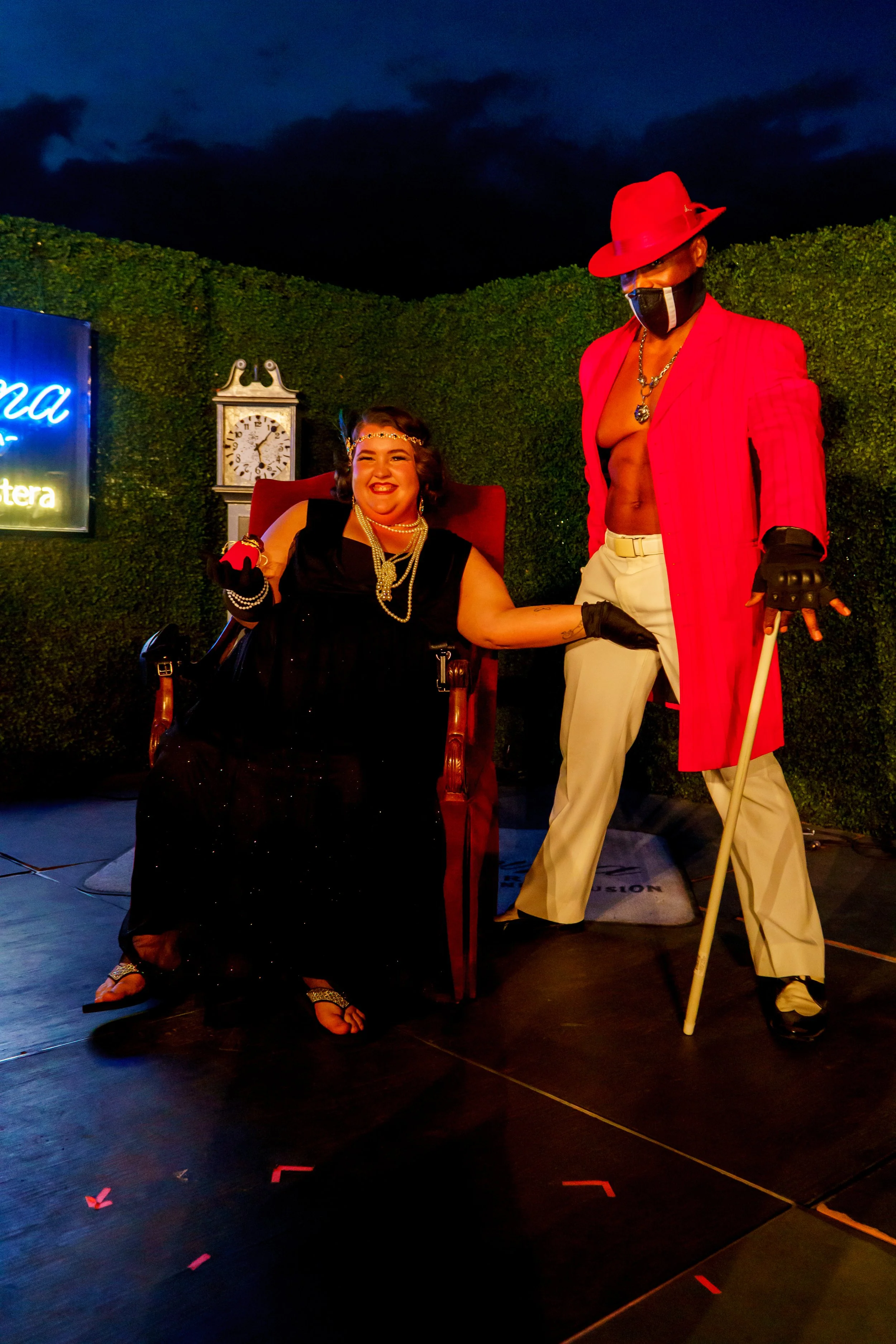 A person in a wheelchair wearing a black dress with pearl jewelry smiling, holding hands with a standing person dressed in a red blazer, white pants, a red hat, a black face mask, and gloves, with a green hedge backdrop and a clock in the background.