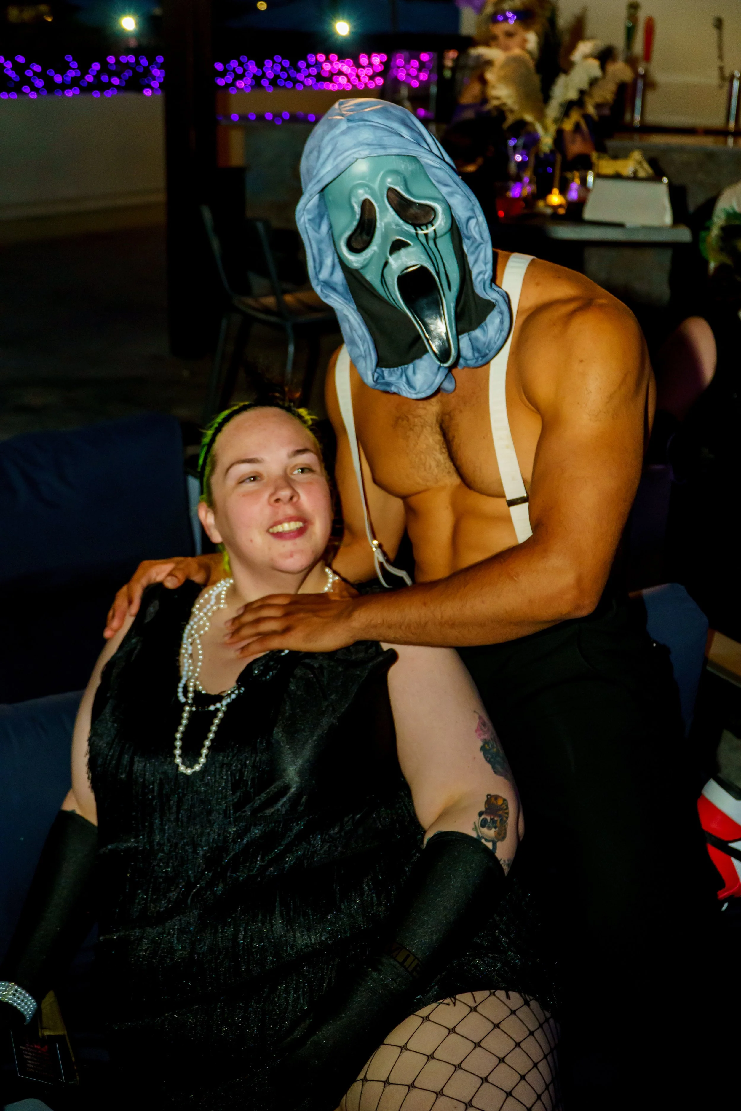 Person wearing a 'Scream' mask with a blue hood, shirtless with suspenders, posing with a woman dressed in a black dress and pearls, sitting on a chair with fishnet stockings in a dimly lit room decorated with purple lights.