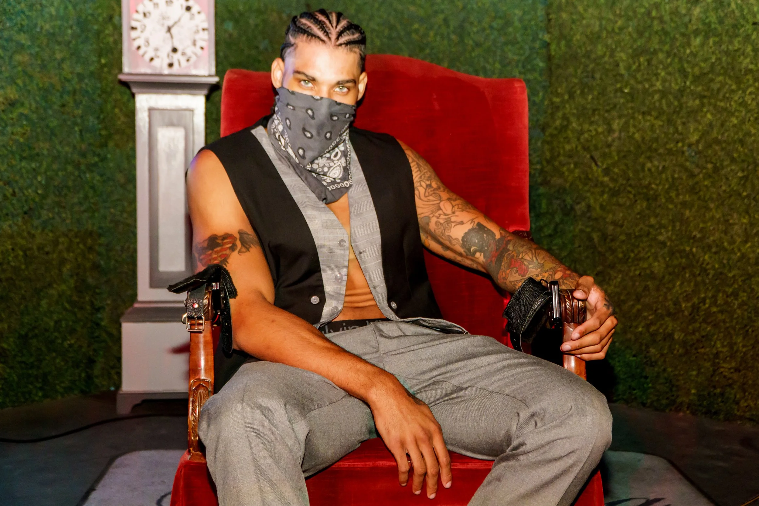 A person with braided hair and tattoos sitting on a red velvet chair, wearing a face mask, sleeveless vest, and gray pants, in front of a green textured background and a tall clock.