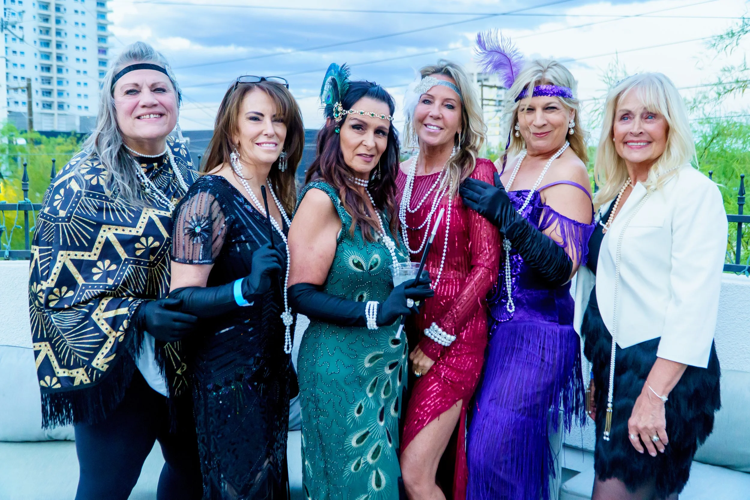 Group of women dressed in vintage 1920s flapper costumes, smiling outdoors with city buildings and cloudy sky in the background.