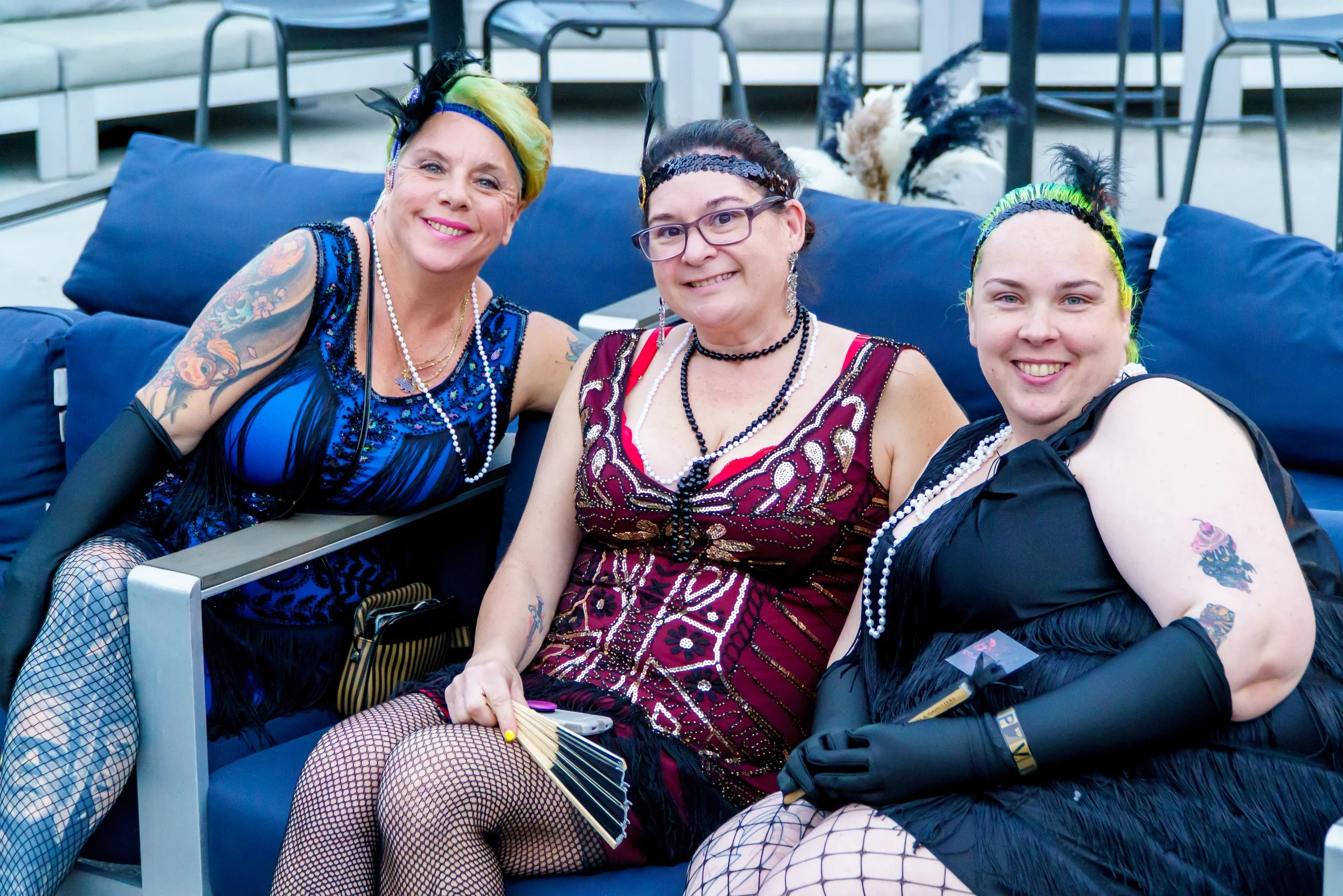 Three women dressed in burlesque or cabaret costumes sitting on a blue couch, smiling at the camera, with a background of chairs and tables.