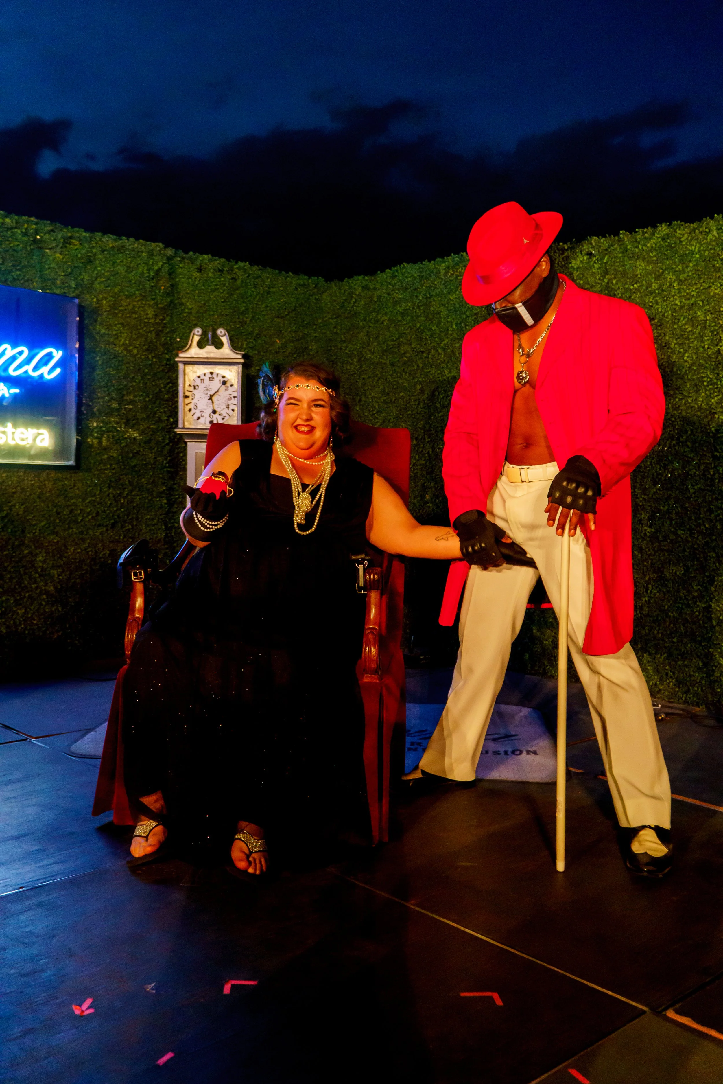 A person dressed as a woman sitting on a red armchair, wearing a black dress, pearl necklace, and a headband, smiling and holding hands with a man dressed as a woman, wearing a red coat, white pants, black gloves, a large red hat, and a black face ma
