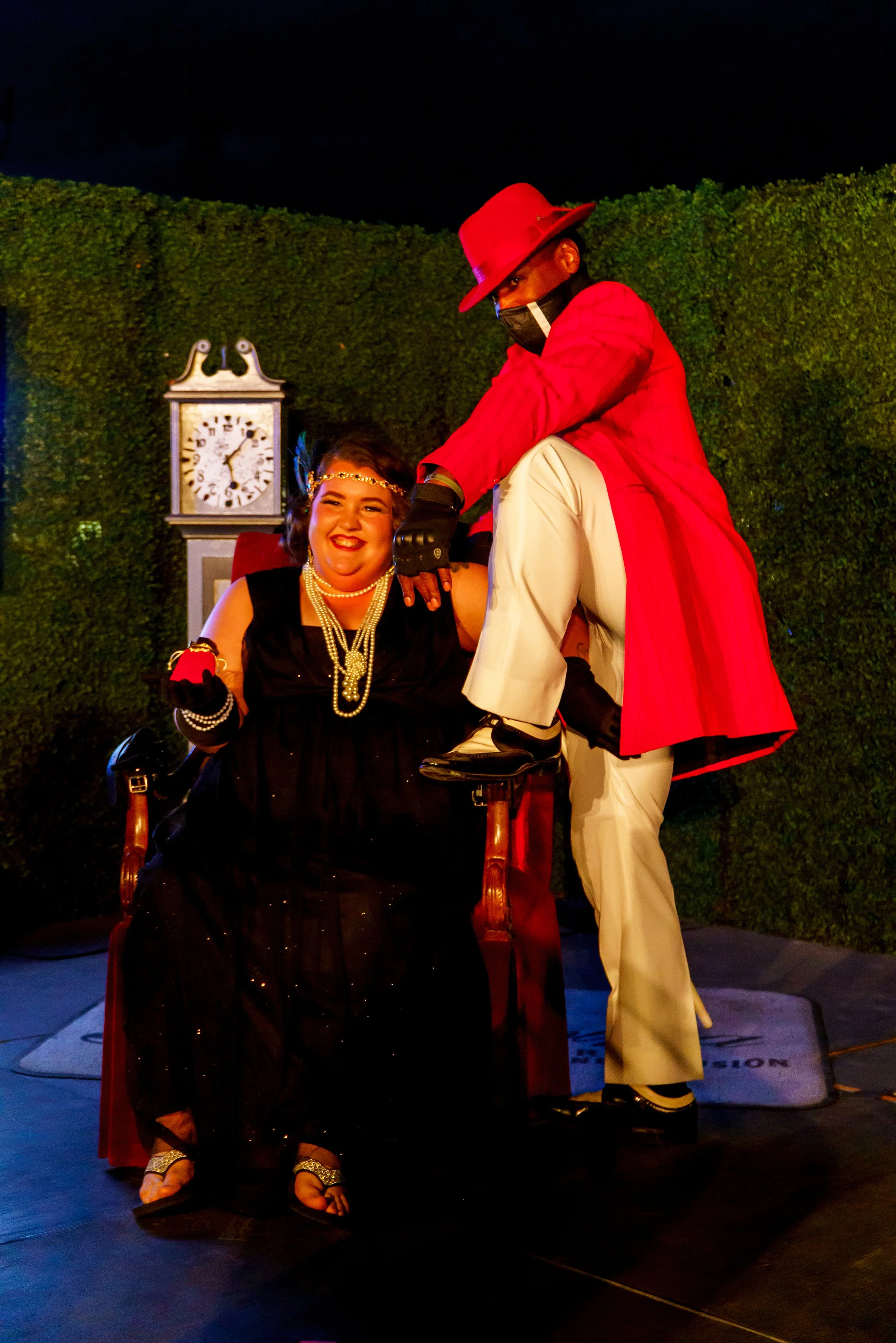 A woman dressed in a black gown and pearl jewelry is sitting in a wheelchair, smiling and holding a red rose. A man dressed in vintage red and white attire with a red hat is kneeling beside her, with his hand on her shoulder, in front of a green hedg