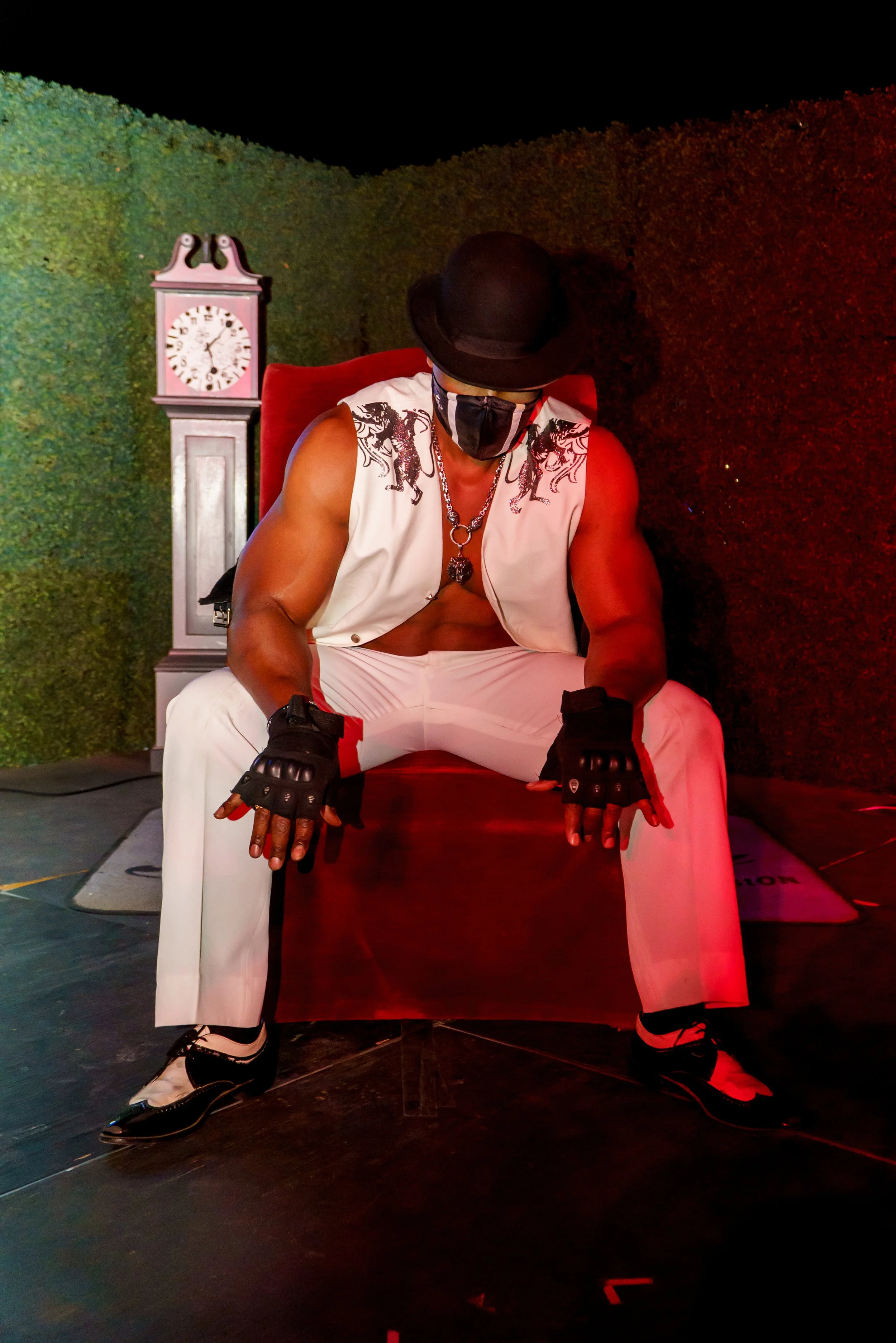 A man sitting on a red chair, wearing a black hat, black mask, white sleeveless shirt with black dragon designs, white pants, and black and white shoes, in front of a green hedge and a vintage pink clock.