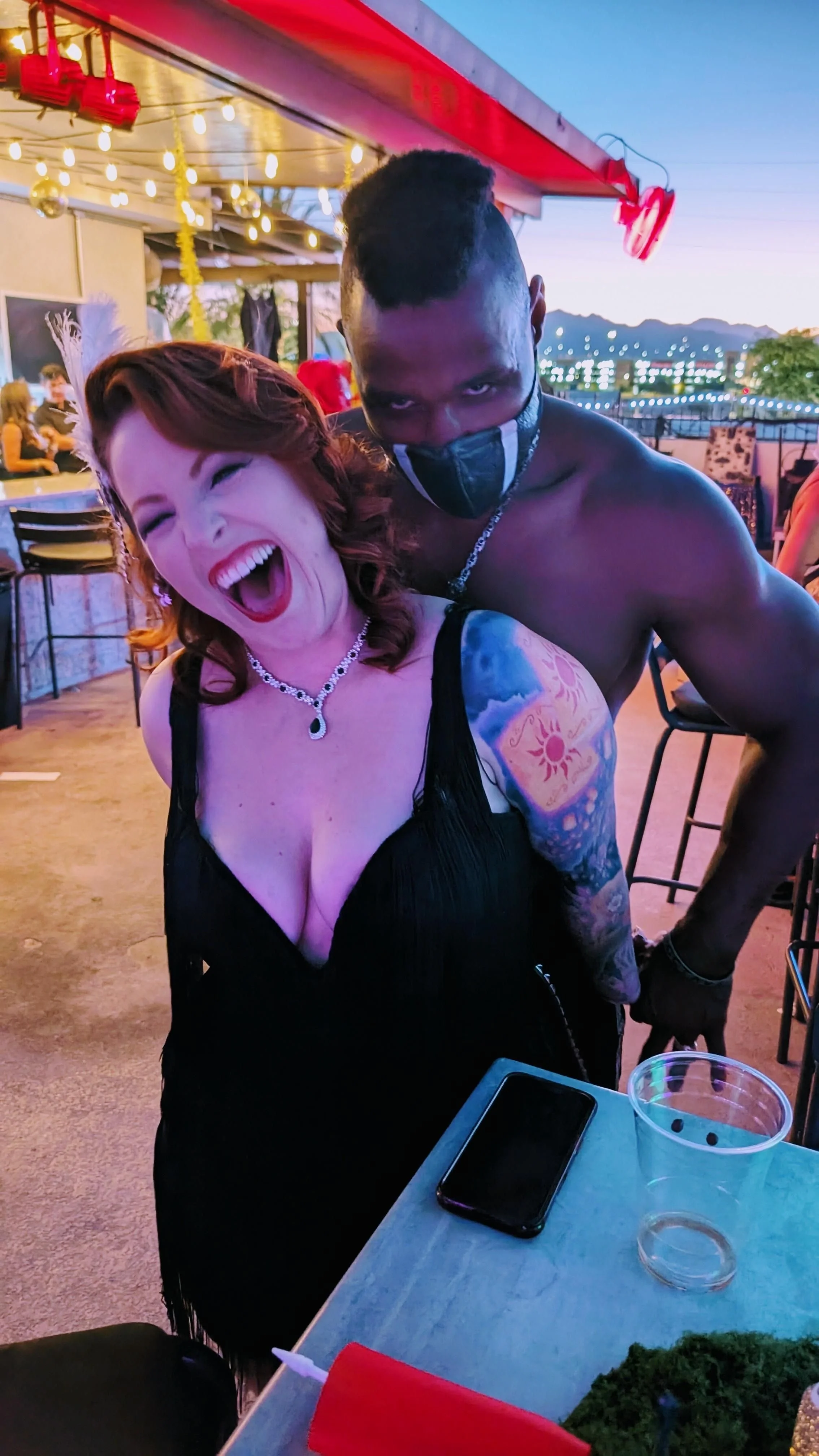 A woman with red hair laughing and a man with dark skin and short hair wearing a black face mask, both at a party or social gathering outdoors during sunset with string lights and mountains in the background.