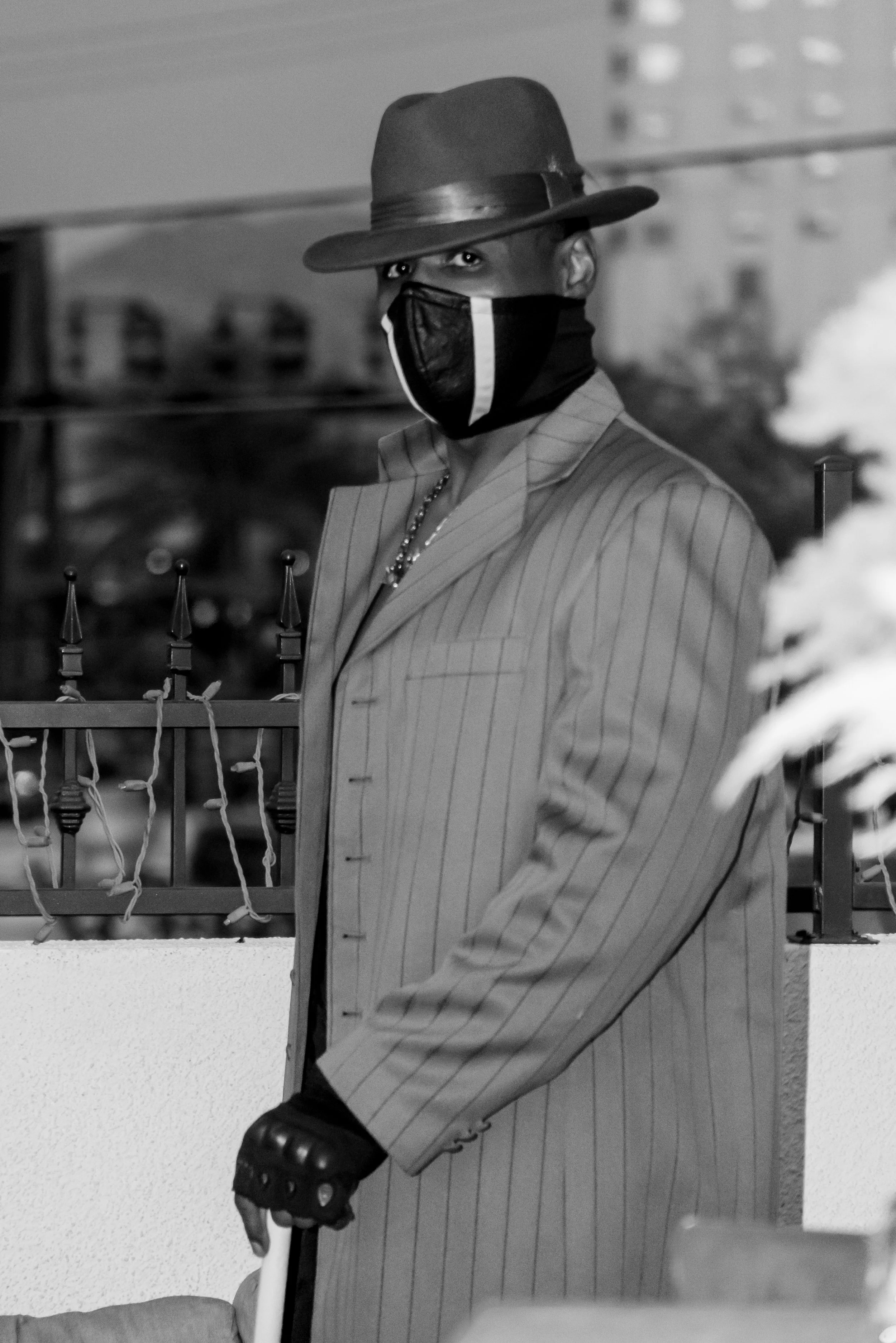 A person wearing a wide-brimmed hat and a face mask looking directly at the camera, dressed in a pinstripe suit.