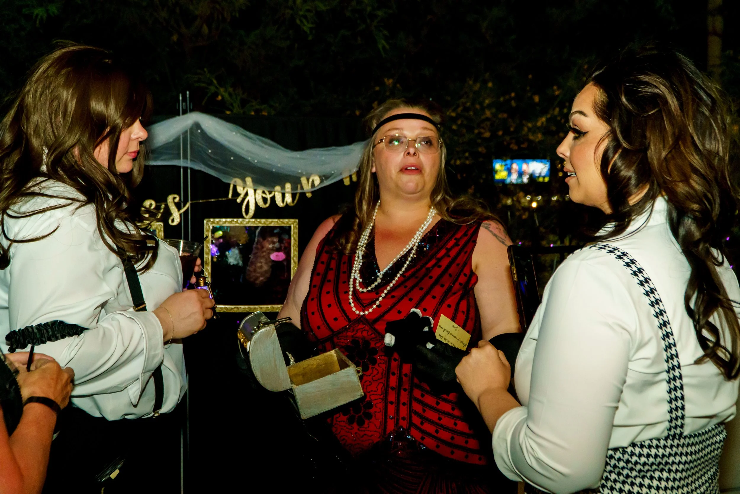 Three women talking at a party, with one in the middle wearing glasses, a red and black dress, and pearls, and the other two women with brunette and curly hair, wearing white tops. There is a sign with gold lettering and a backdrop with lights.