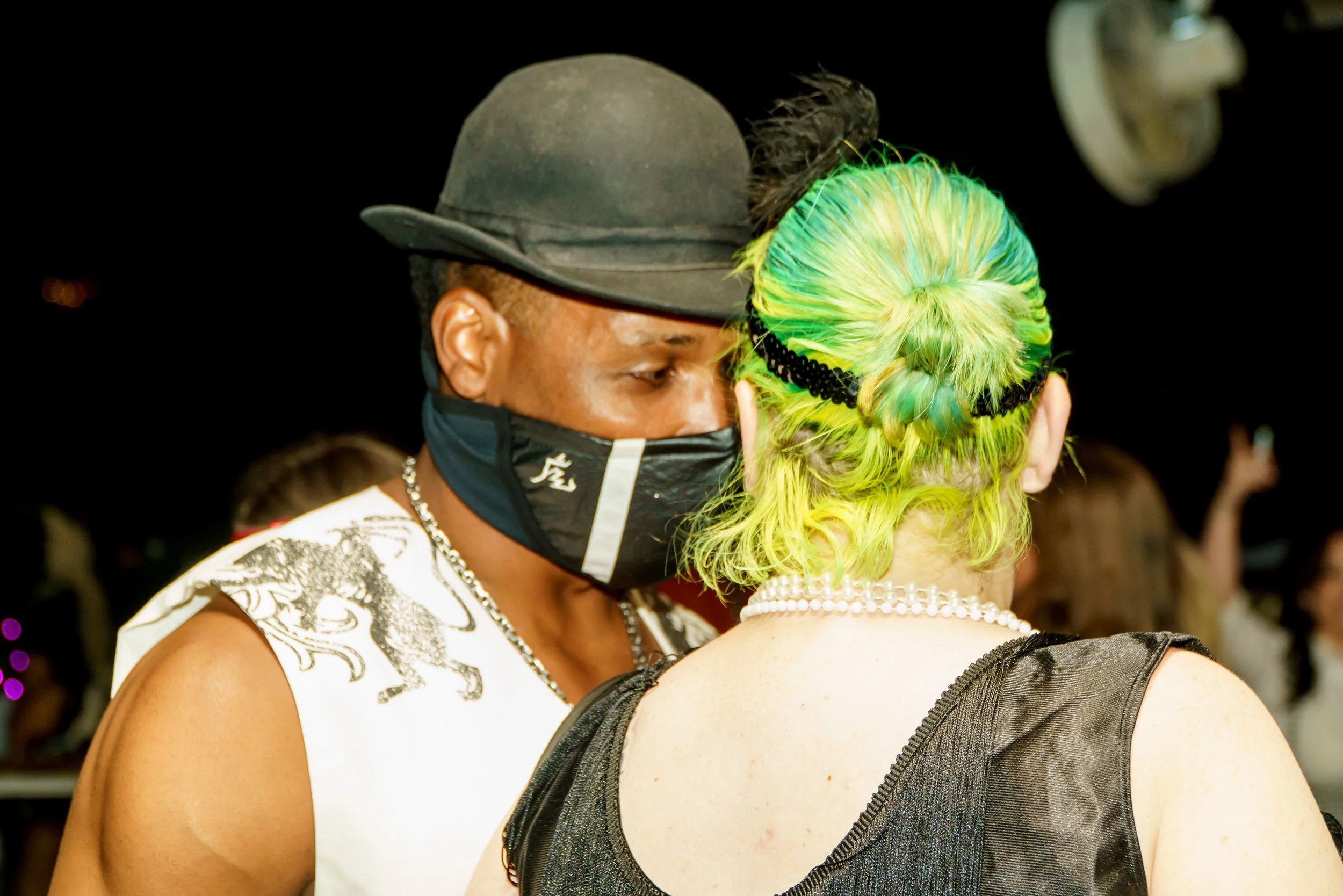 A man wearing a black hat and face mask with a woman with green and yellow hair and a pearl necklace, embracing at night with black background.