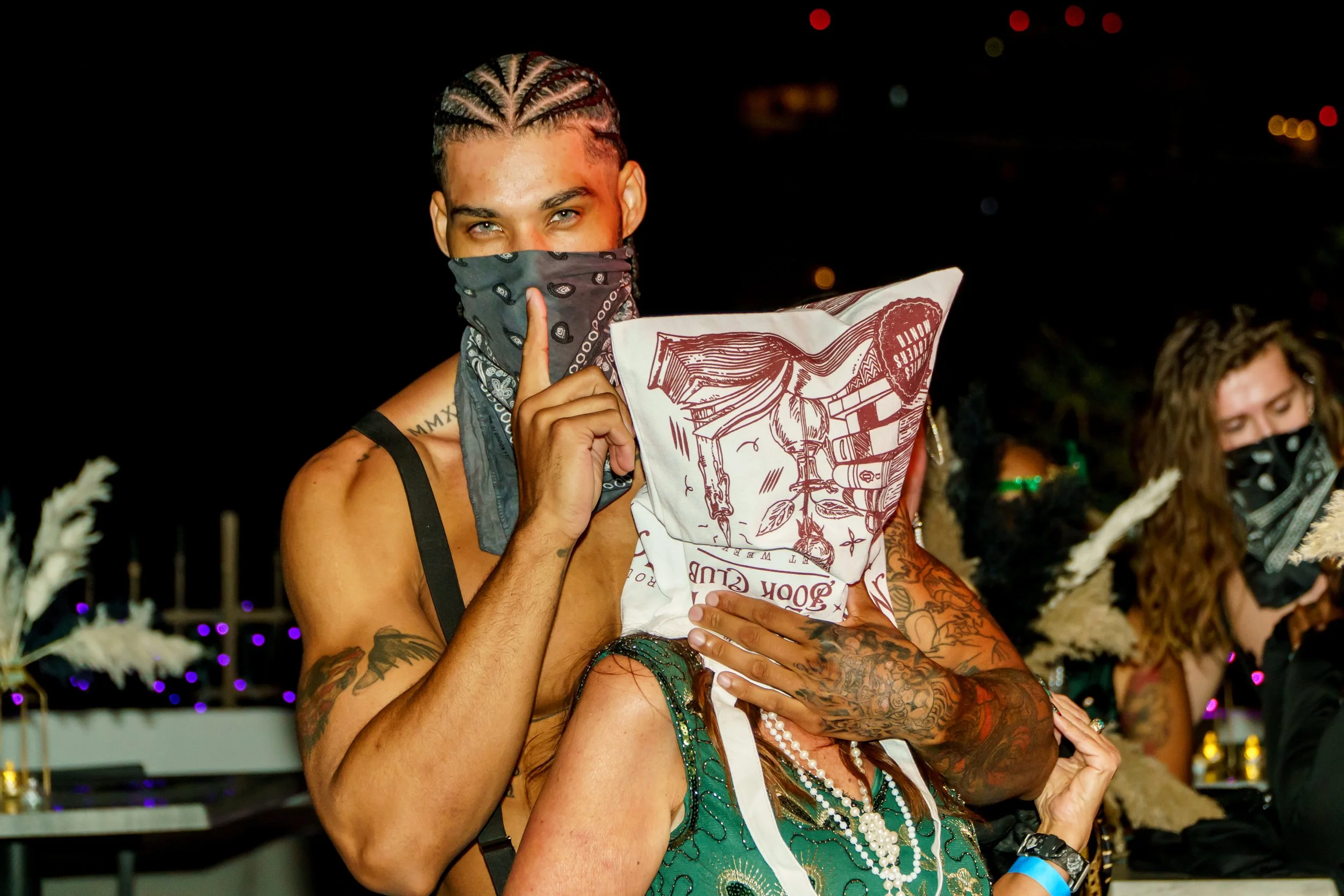 A man with braided hair and a bandana covering his face is holding a pillow while hugging a woman wearing a green dress and pearl necklaces at an outdoor event at night.