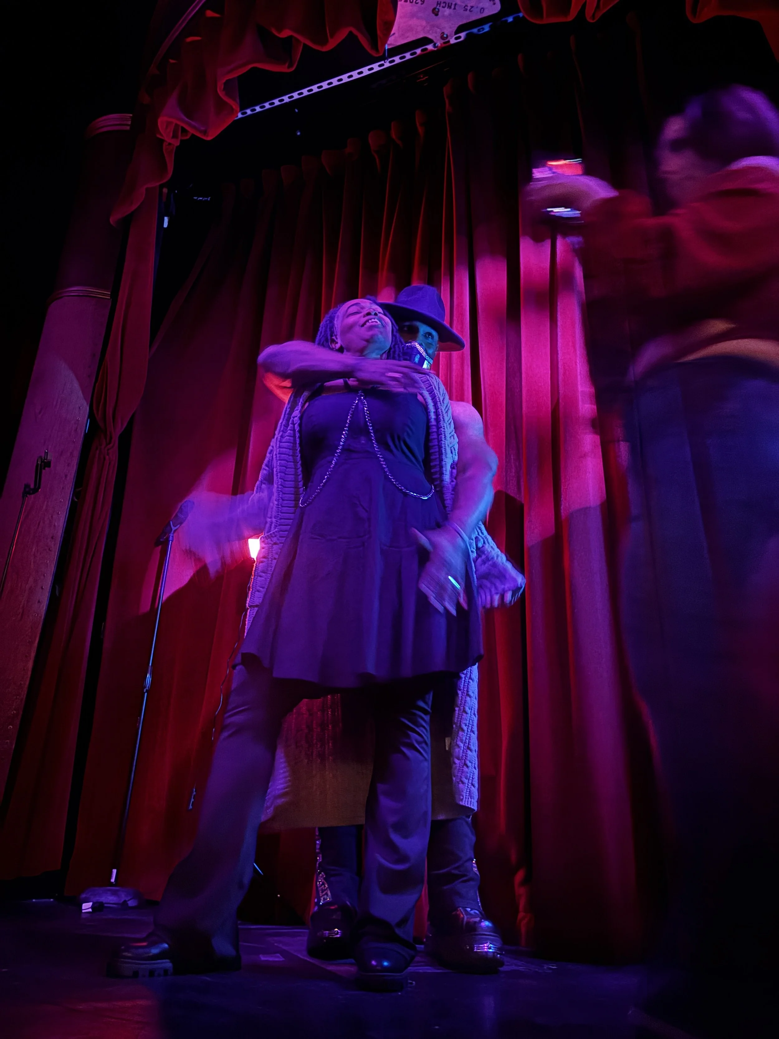 Two performers standing on a stage with red curtains, illuminated by purple and pink stage lights.
