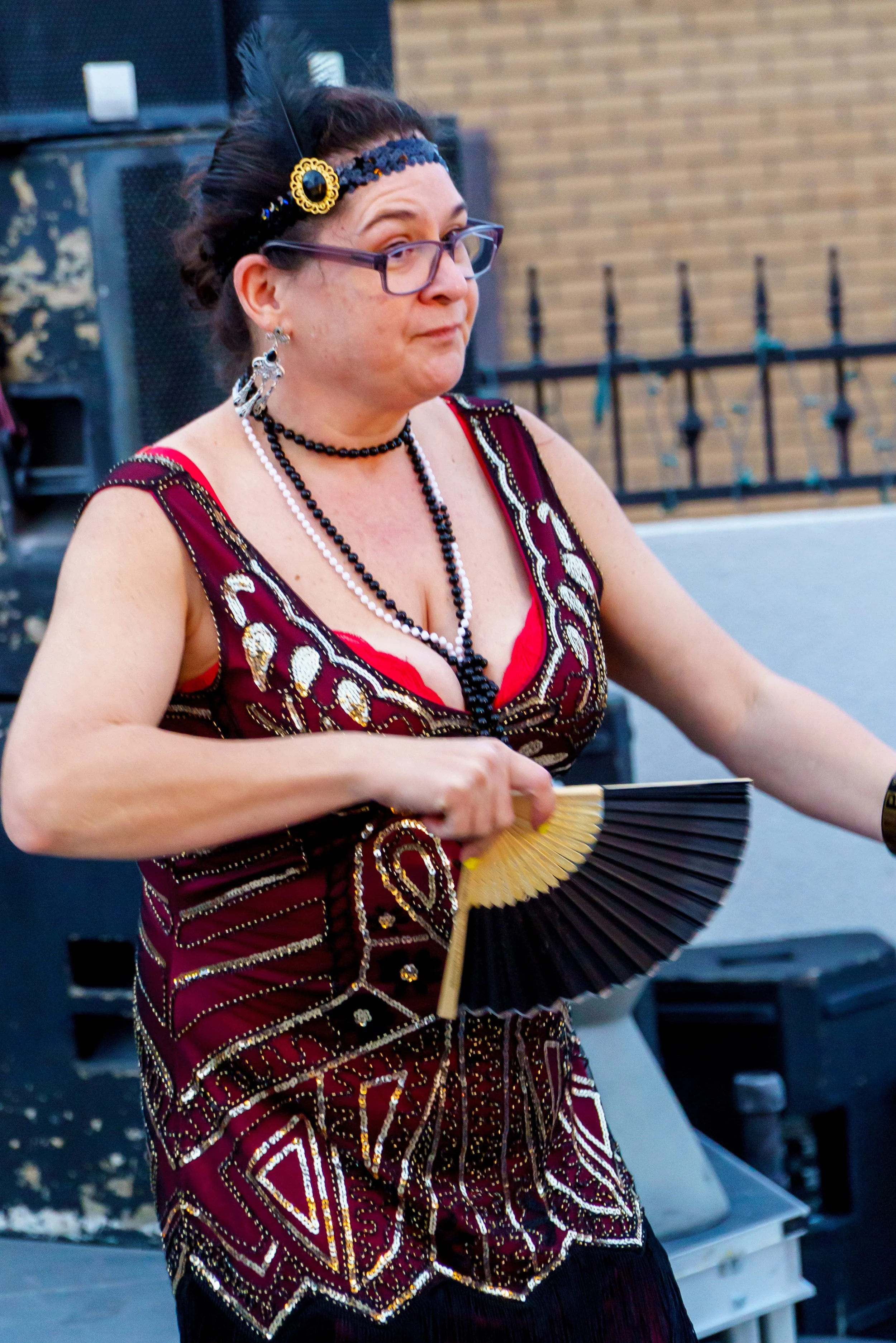A woman with dark hair wearing glasses and ornate jewelry, including earrings, a headband, and necklaces, holding a black and gold folding fan in an outdoor setting.