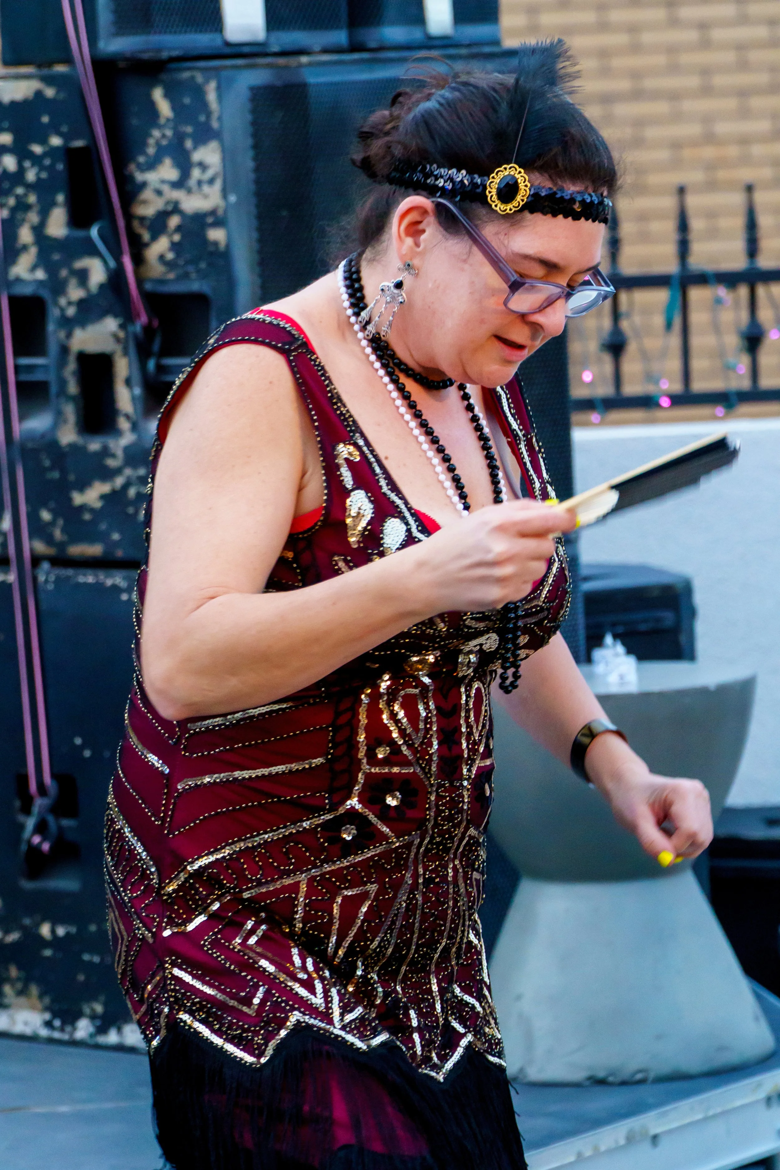 A woman with dark hair and glasses, dressed in a red and gold sequined dress, wearing large earrings and a beaded necklace, looking at her phone at an outdoor event.